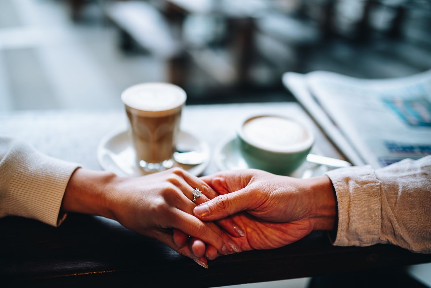 Two hands reach across a table to hold each other, showing her engagement ring, while coffee cups sit nearby in Melbourne photo session