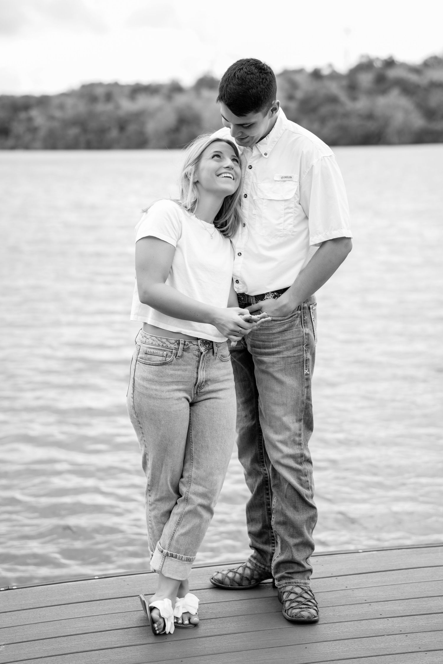 Black and white photo of a couple embracing on a dock by a lake, woman smiling up at man in casual jeans and shirts.