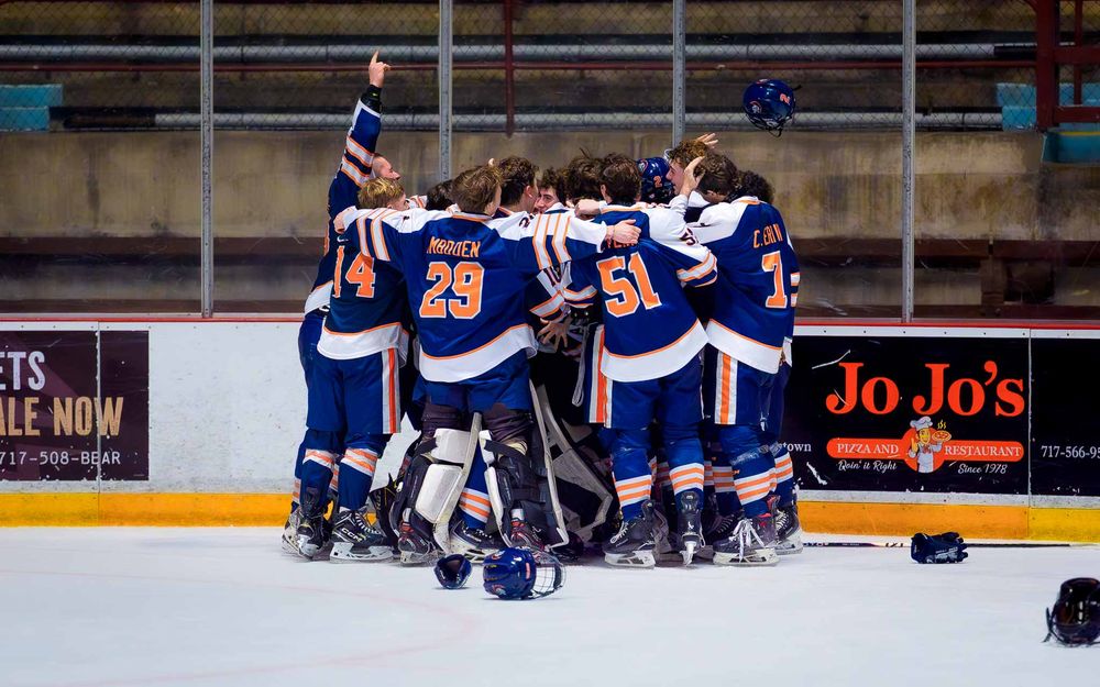 Trojans hockey team huddles at center ice, arms raised in celebration after winning the Bears Cup by SportsFox Photography, Hershey PA.