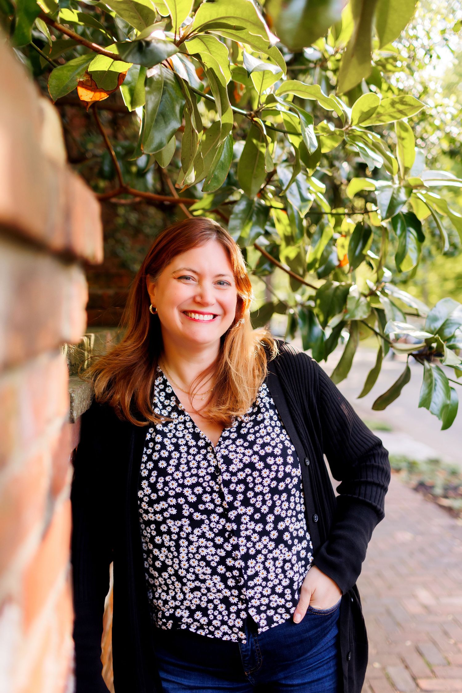 A person wearing a floral print blouse and black cardigan sits outdoors among green foliage and brick walls.
