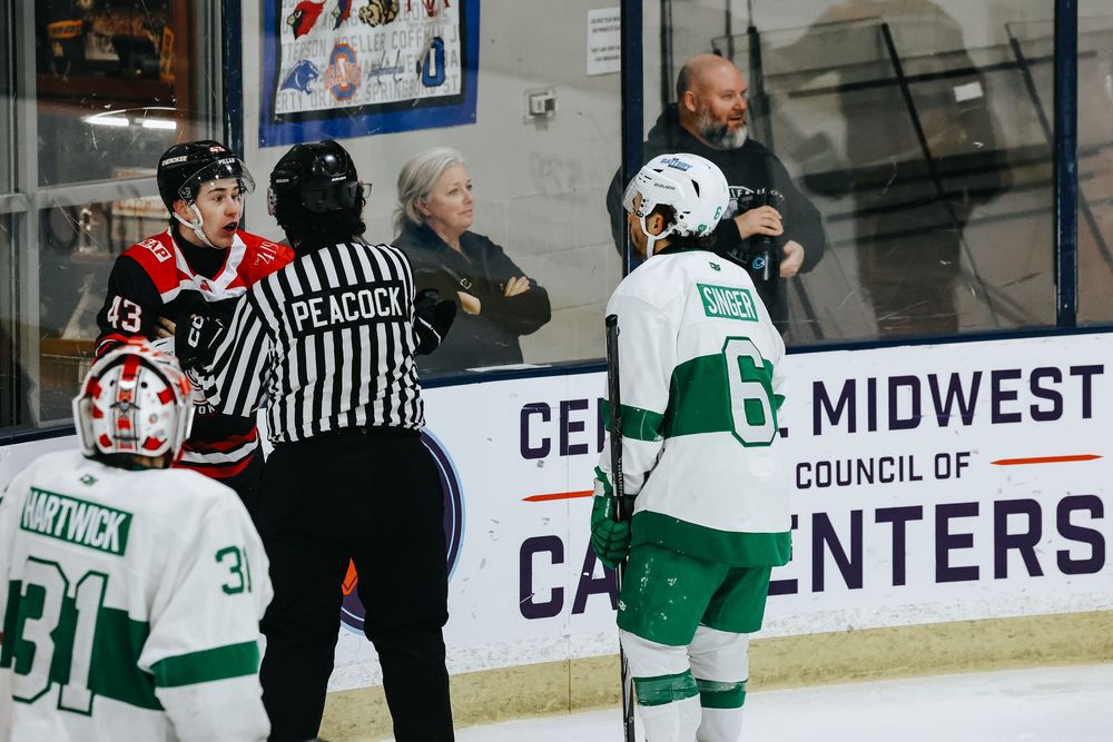 Hockey players and officials gather around the boards during a game with Midwest Carpenters signage visible in background.