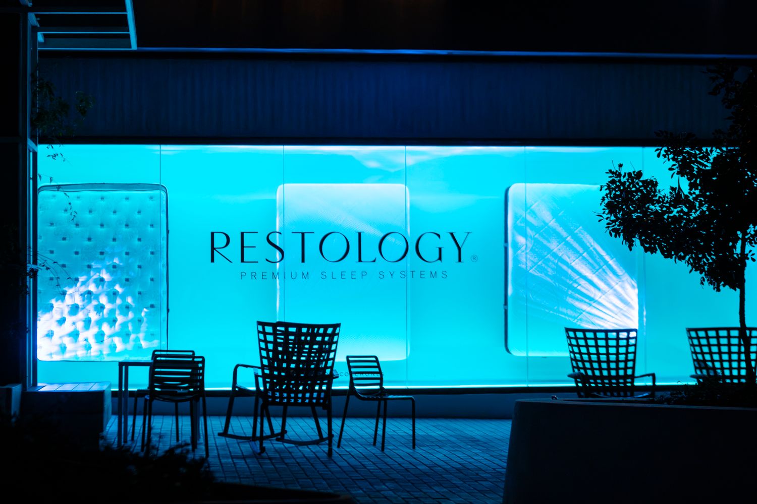 Restology spa exterior glowing in blue neon light with chairs and decorative panels at night.
