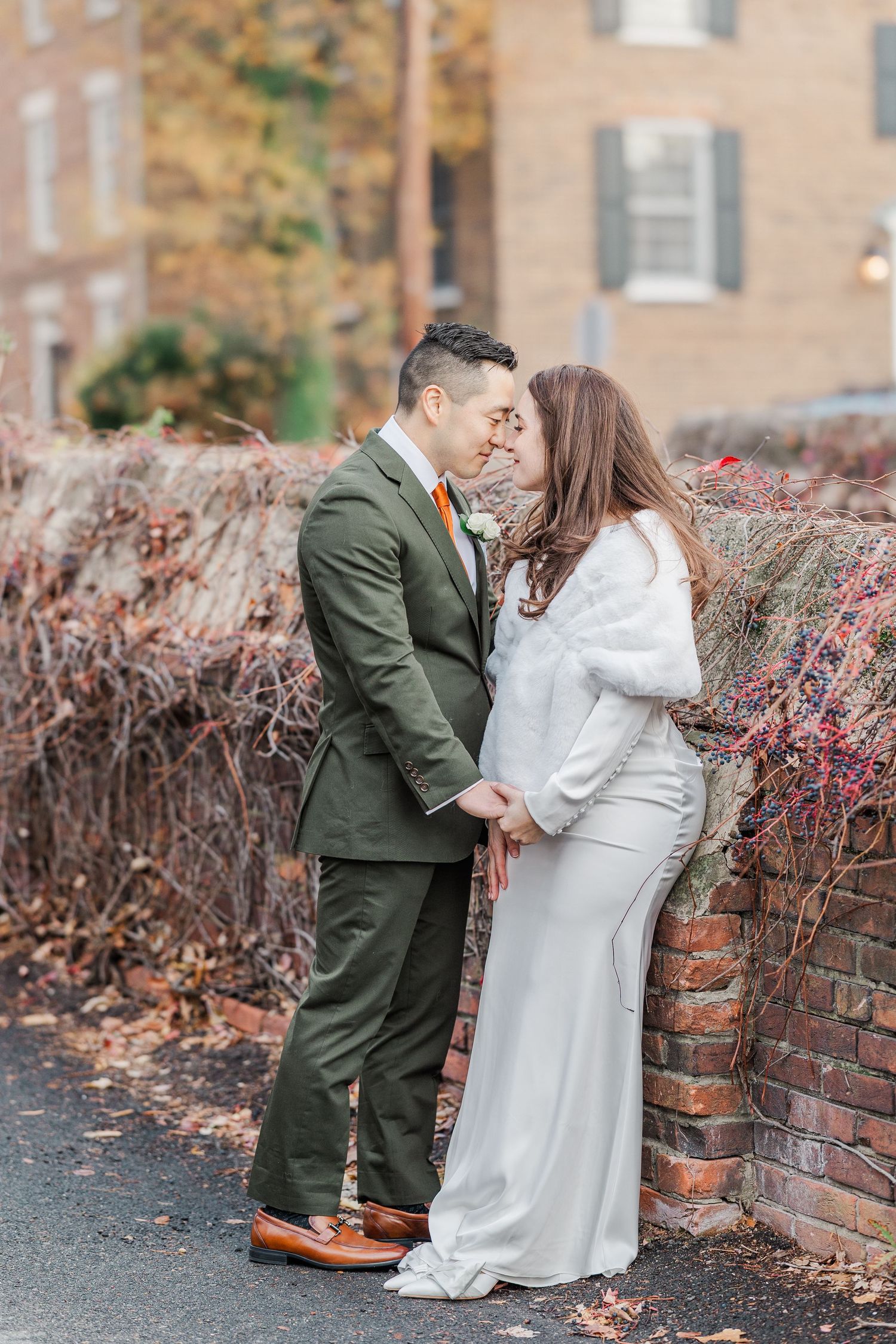 A couple in wedding attire share a romantic moment against a brick wall with autumn foliage in the background.
