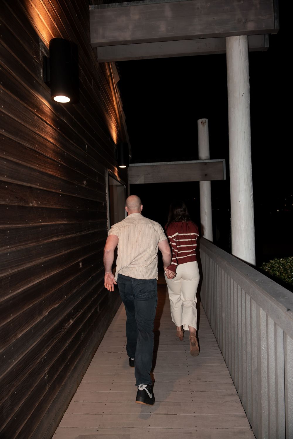 Two people walking down a covered outdoor walkway with wooden walls and white columns at night.