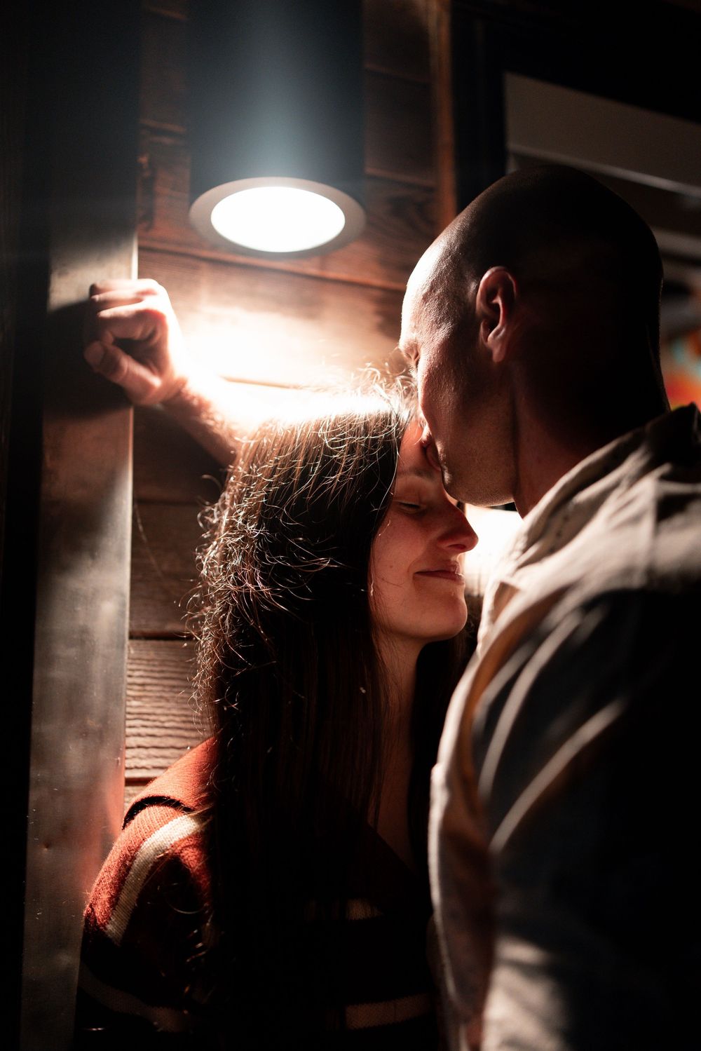 A couple shares an intimate moment in dramatic lighting as they lean in close to each other in a dimly lit setting.