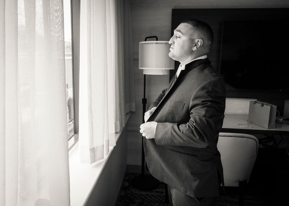 Groom in black suit adjusting tie while gazing out hotel window in black and white wedding photo.