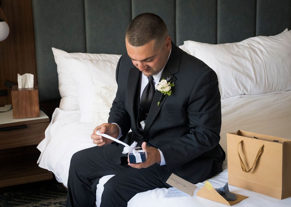 Groom in black suit with white boutonniere opening a gift box while sitting on a hotel bed.