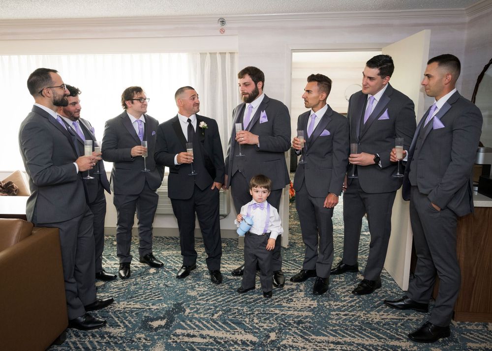 Groomsmen in gray suits with purple ties gather with a young ring bearer in a hotel room before a wedding ceremony.
