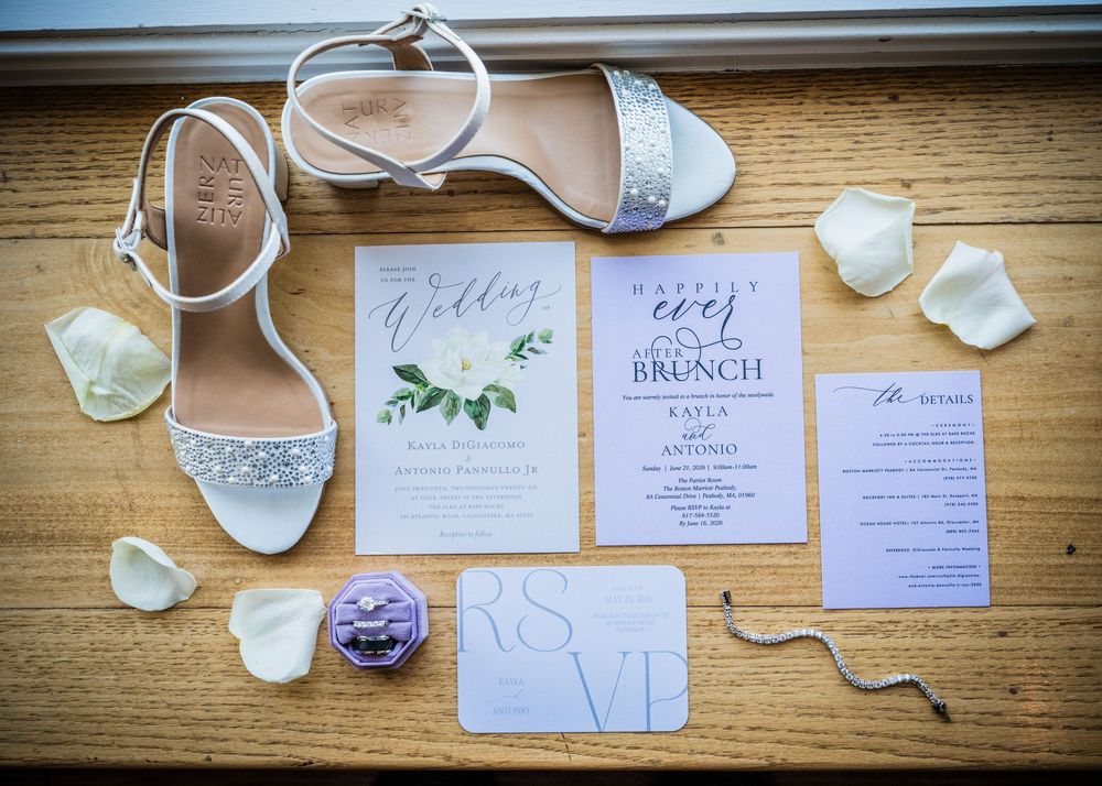 Elegant wedding invitation suite with white heels, rose petals, and purple ring box on wooden surface.