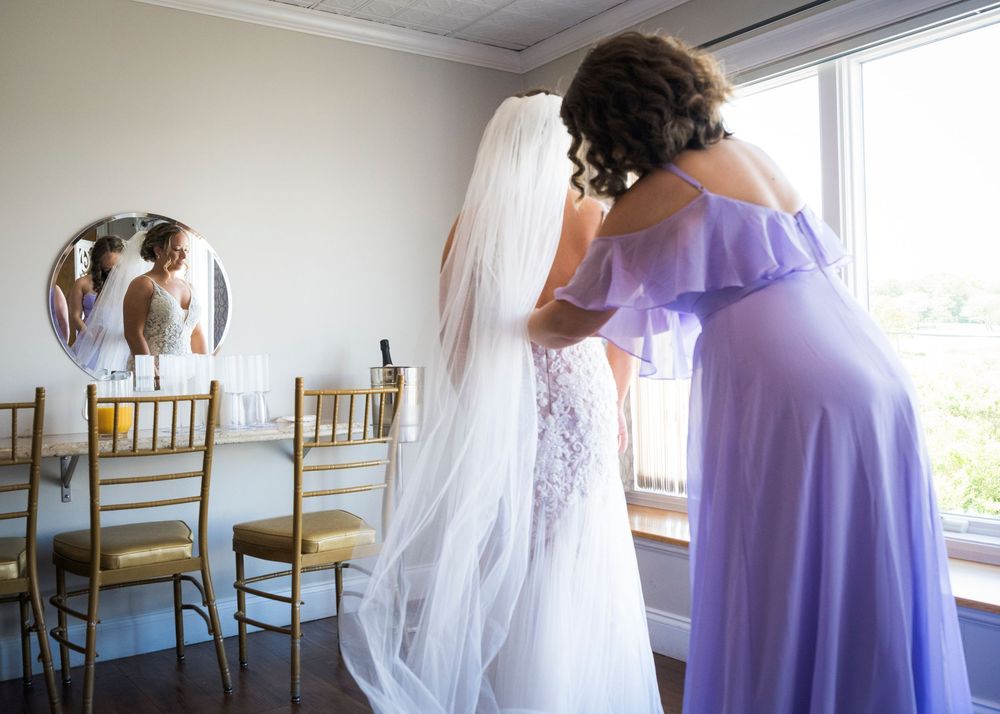 Bridesmaid in lavender gown helping bride in white wedding dress and veil near a window in bridal suite.