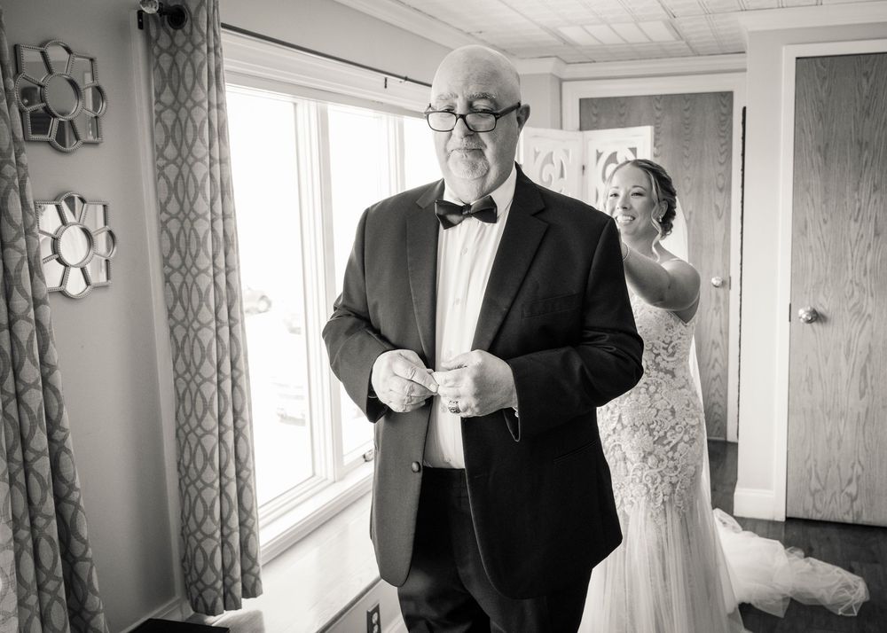 Father of the bride adjusts his bow tie as his daughter in a lace wedding gown stands behind him in black and white photo.