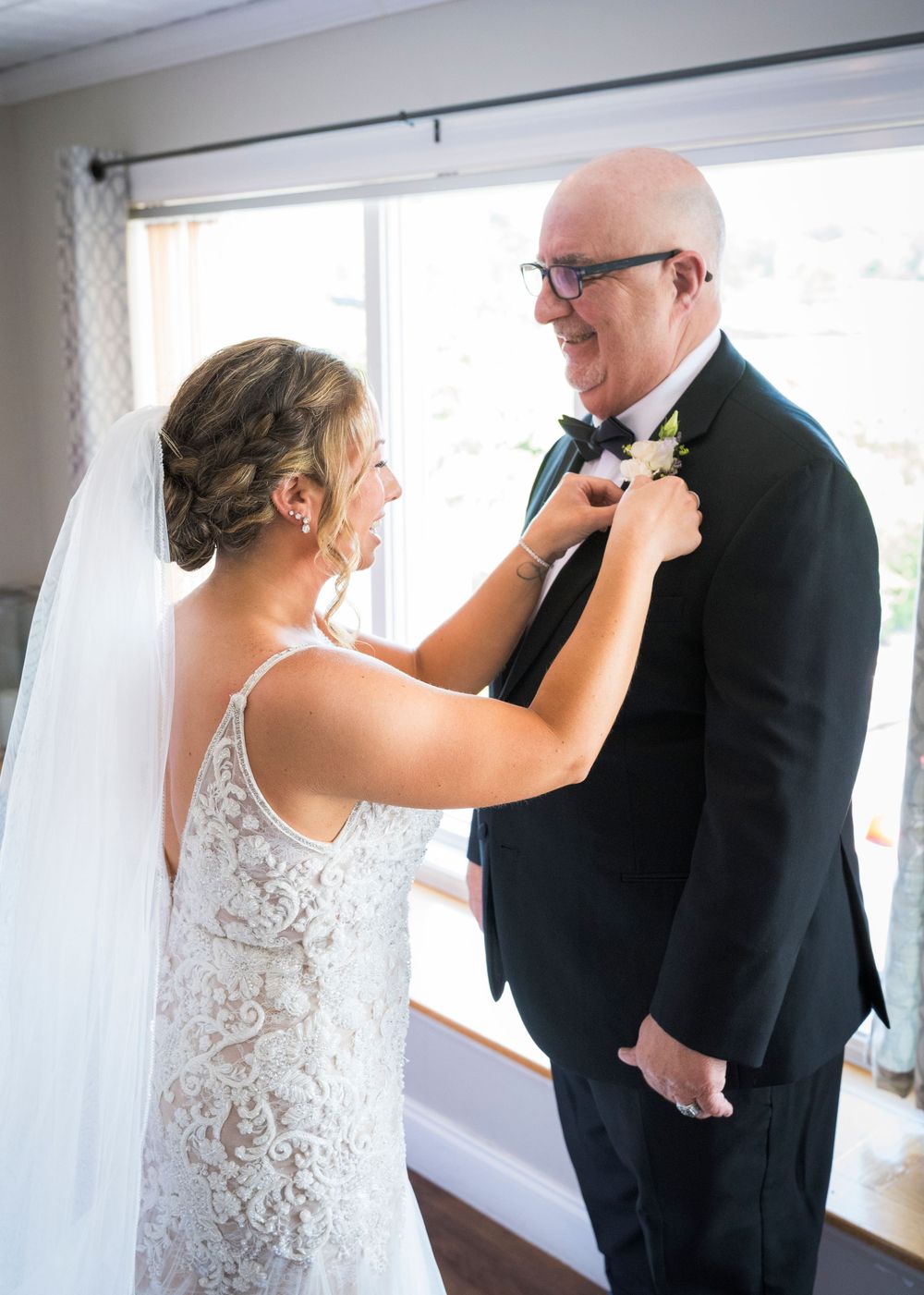 Bride in lace gown adjusting boutonniere on fathers tuxedo lapel by bright window before wedding ceremony.