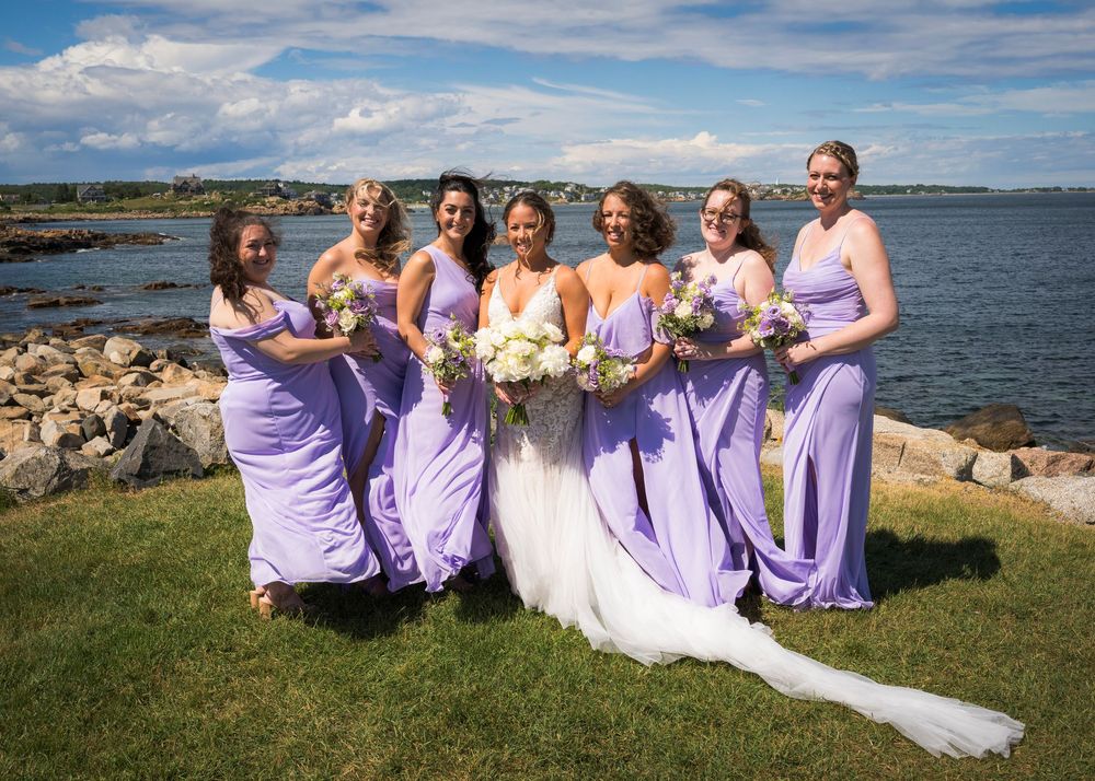Bride with six bridesmaids in lavender gowns holding bouquets by rocky New England coastline.