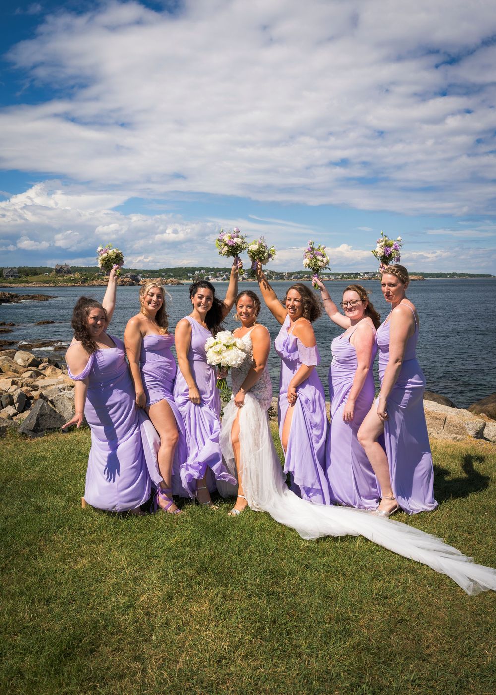 Bride poses with six bridesmaids in lavender gowns holding bouquets by scenic ocean waterfront.