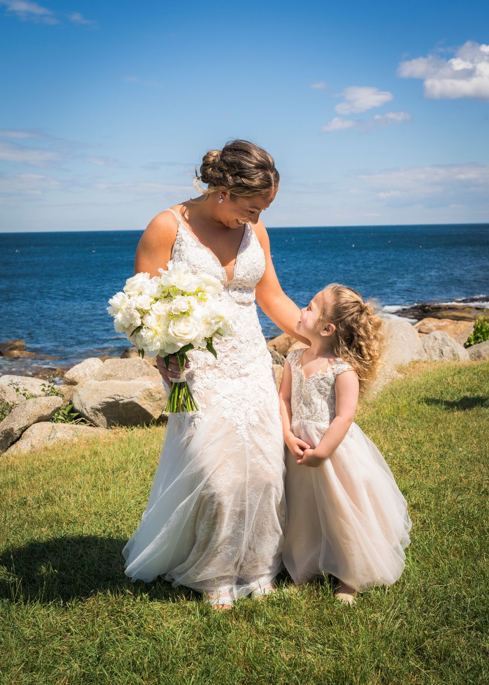 Bride in lace wedding gown holds white bouquet while sharing a tender moment with flower girl by the ocean.