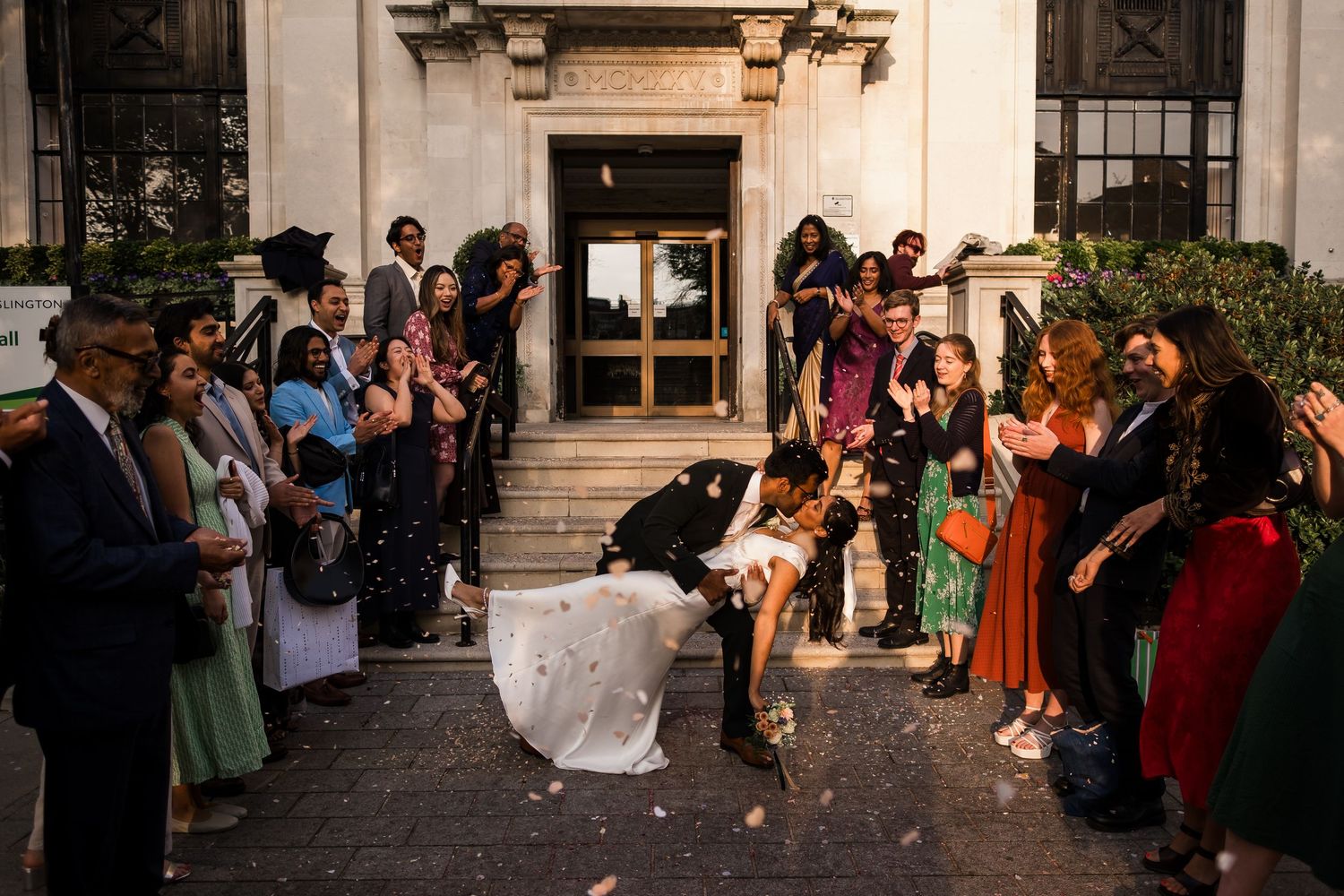 Wedding guests throw confetti on newlyweds outside historic building entrance during daytime celebration.