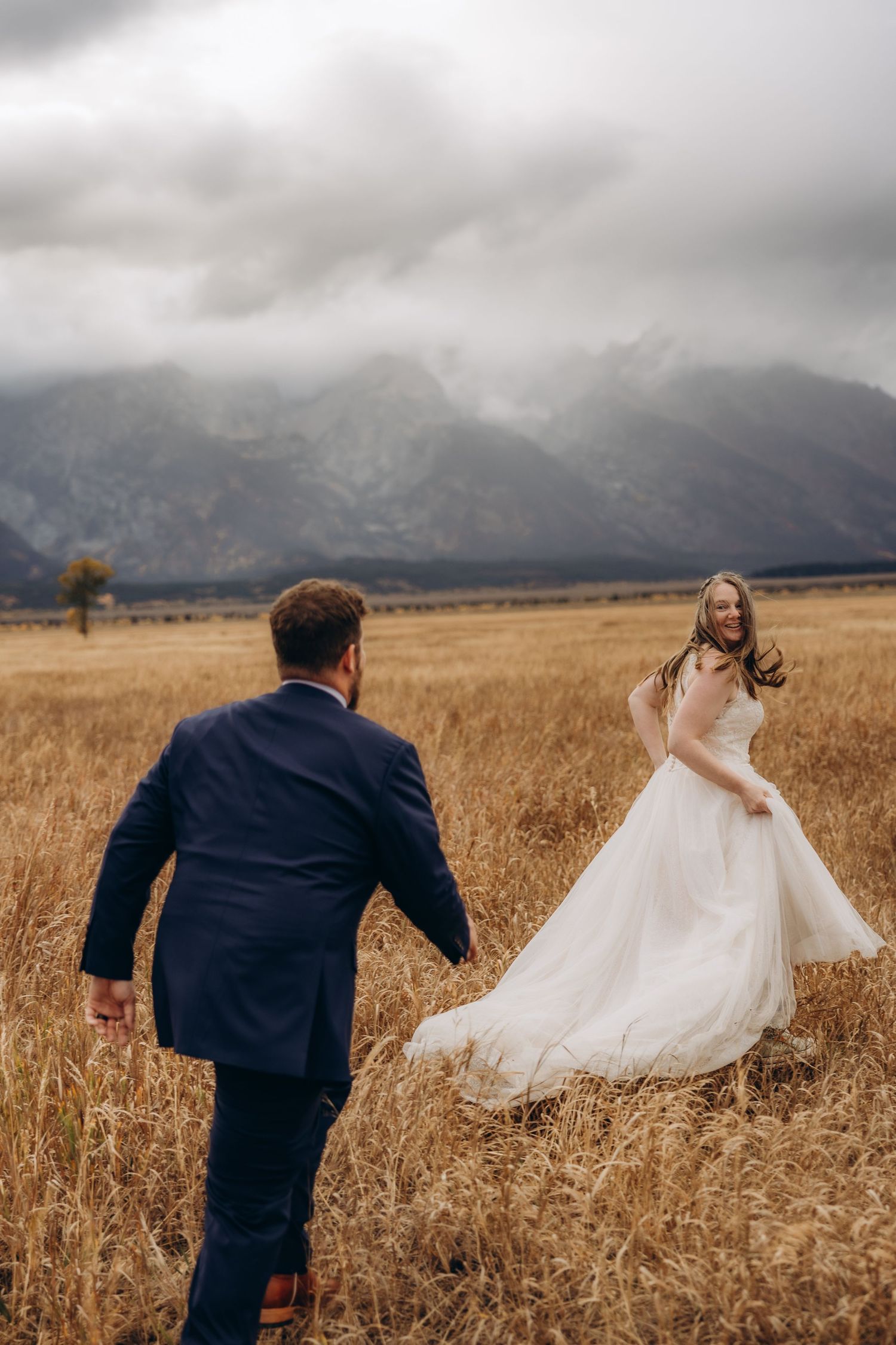 A couple in wedding attire dances in a golden field with dramatic mountain peaks and moody clouds in the background.