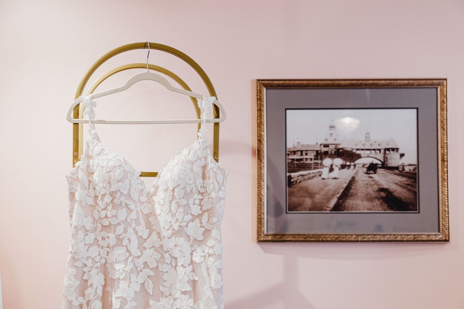 A delicate white lace wedding dress hangs from a gold hanger next to a vintage framed photograph on a pale pink wall.