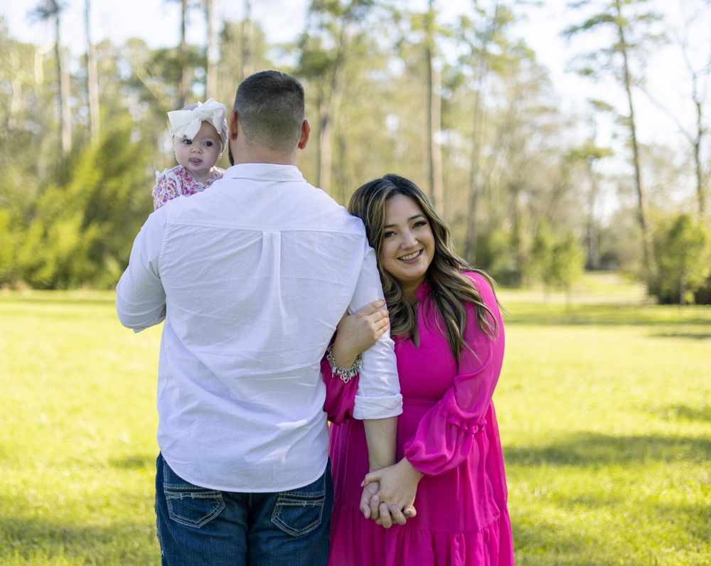 A family in coordinated outfits poses together on a sunny day in a grassy park with tall trees in the background.