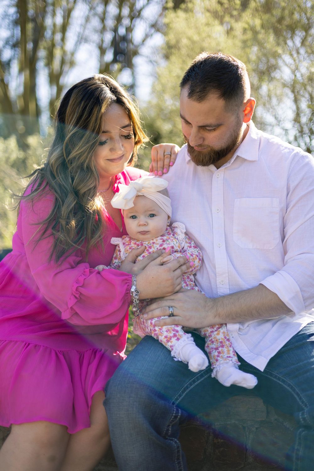 A couple in pink and white outfits sits outdoors with their baby in a floral outfit on a sunny day.
