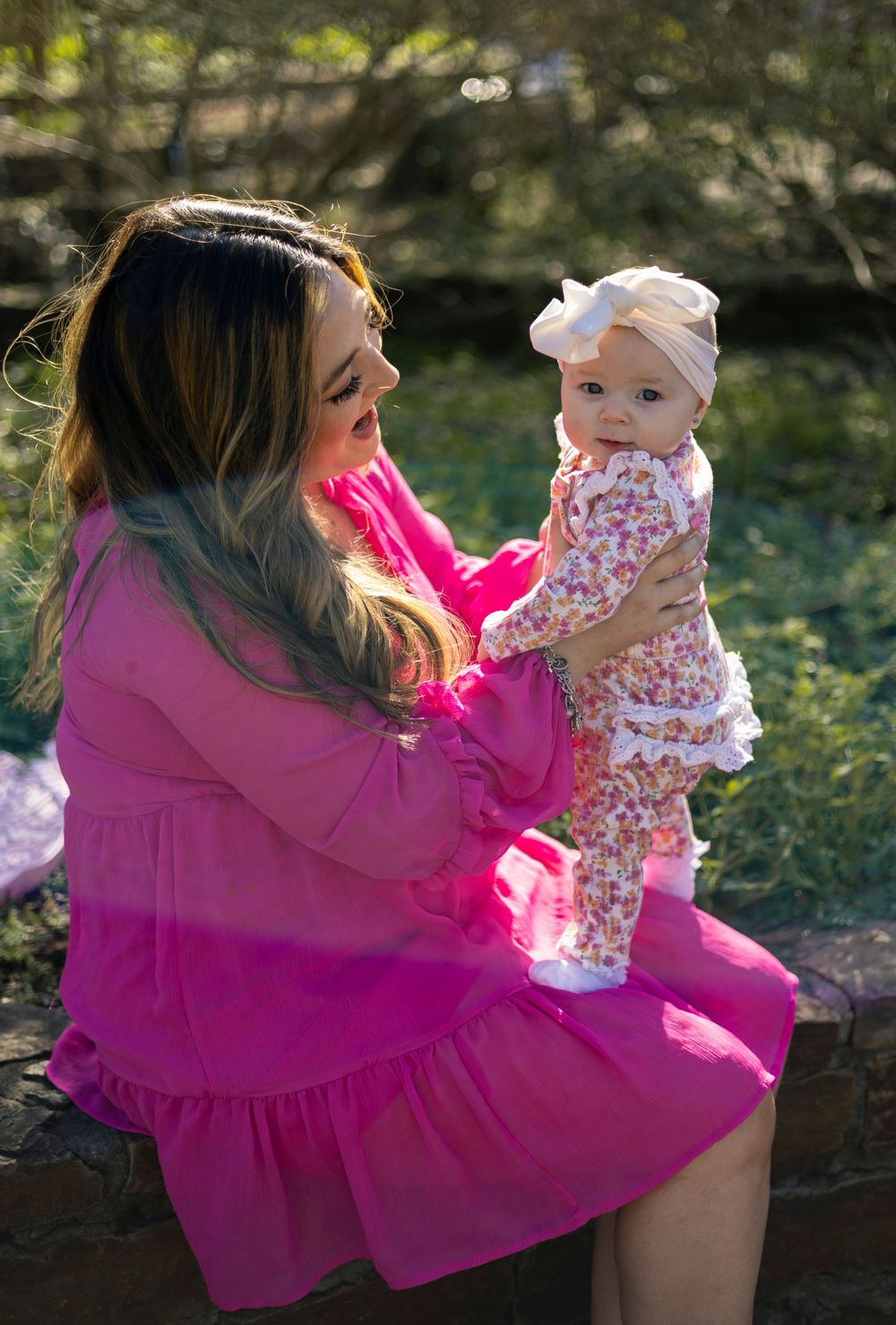 A mother in a bright pink dress lifts and plays with her baby outdoors in a natural setting during golden hour.