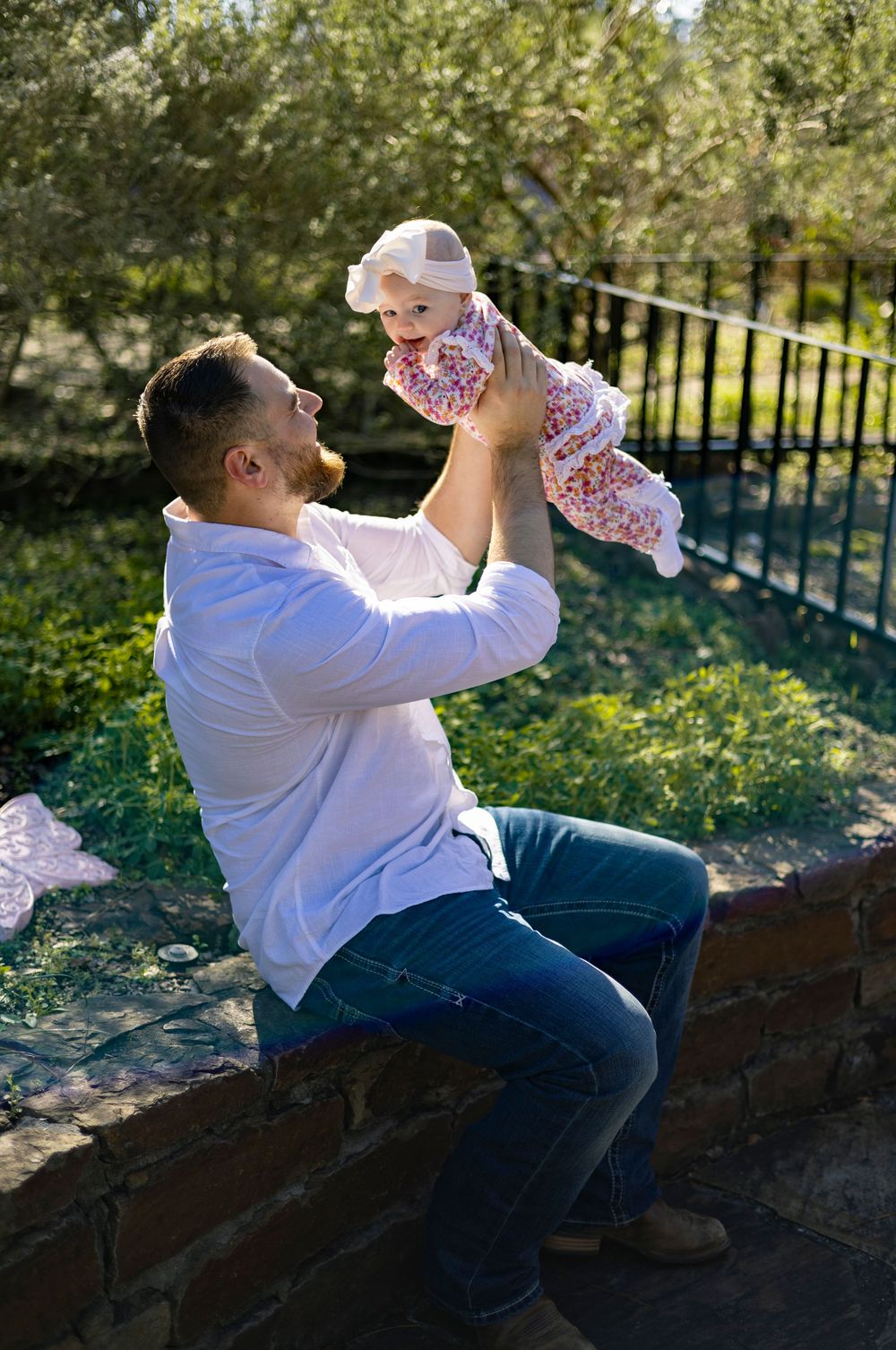 Person in white shirt and jeans sitting on brick wall drinks from bottle on sunny day in park setting.