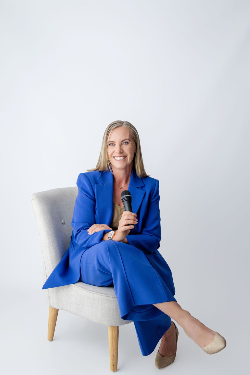 A woman in a vibrant blue blazer sits casually in a white chair holding a microphone, Kirsten Graham Photography