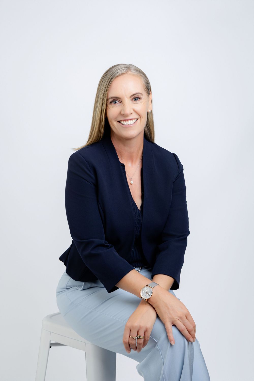 Professional business portraits of woman in a navy blazer and light blue pants seated against white background.