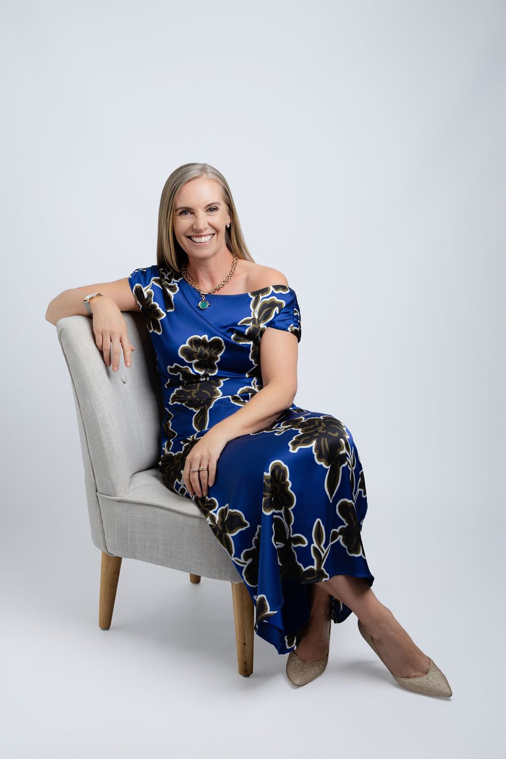 Woman in blue floral dress poses elegantly on a gray armchair during a professional photoshoot.