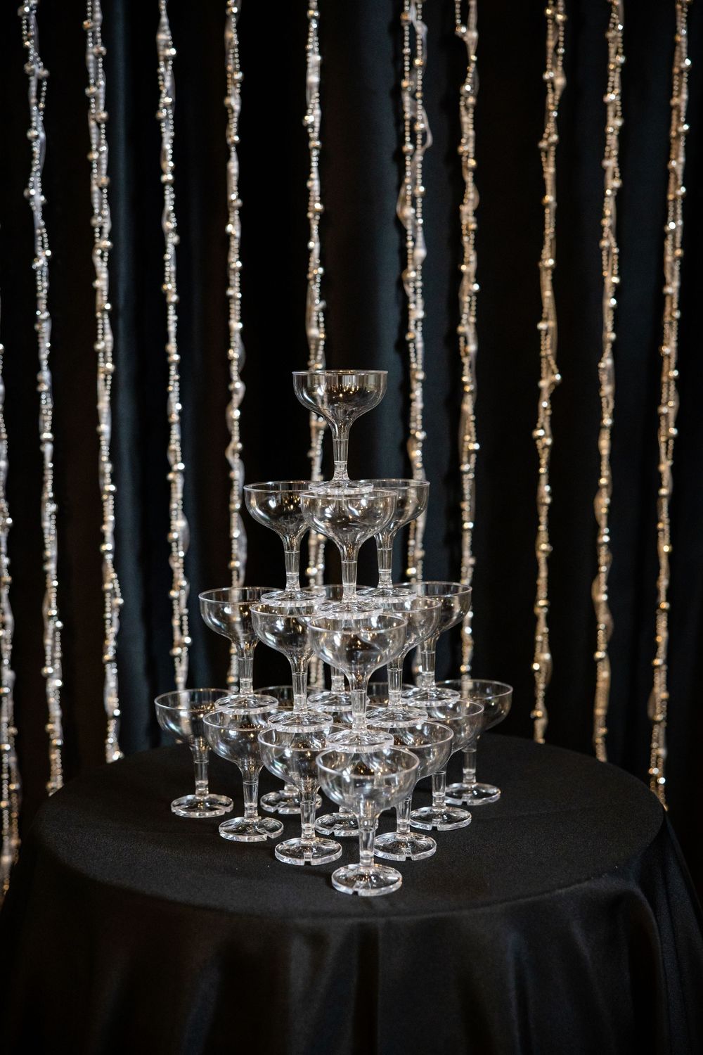 Crystal champagne glasses arranged in a tower formation against a dark background with hanging string lights.