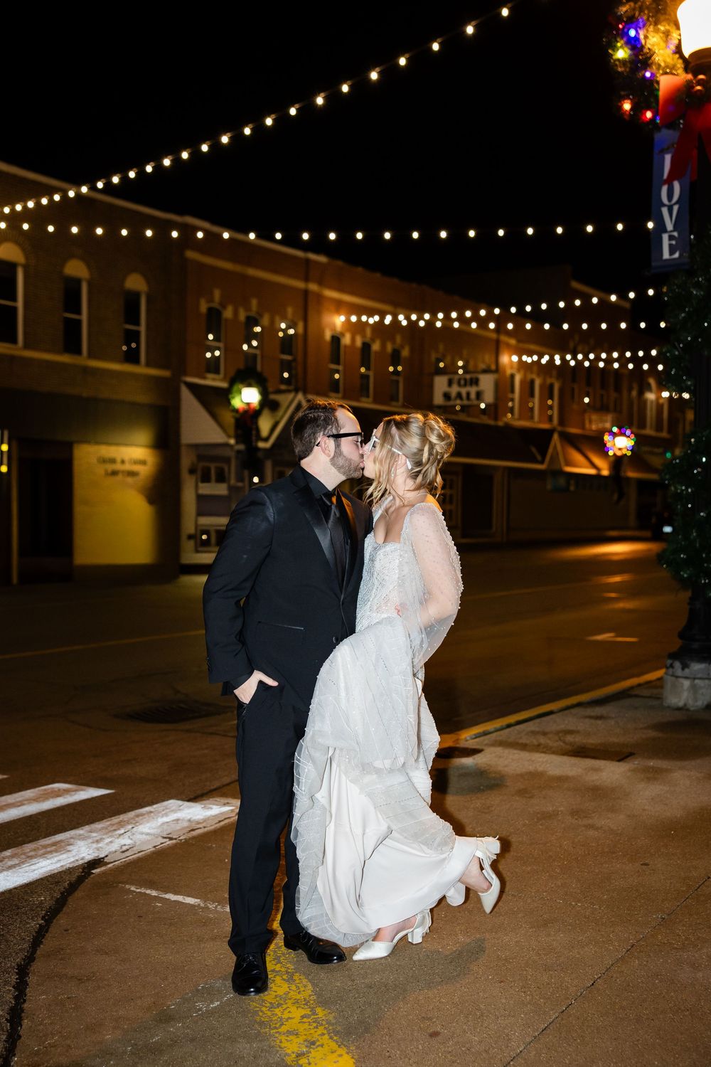 A romantic nighttime street scene with string lights overhead as a couple shares a tender moment.