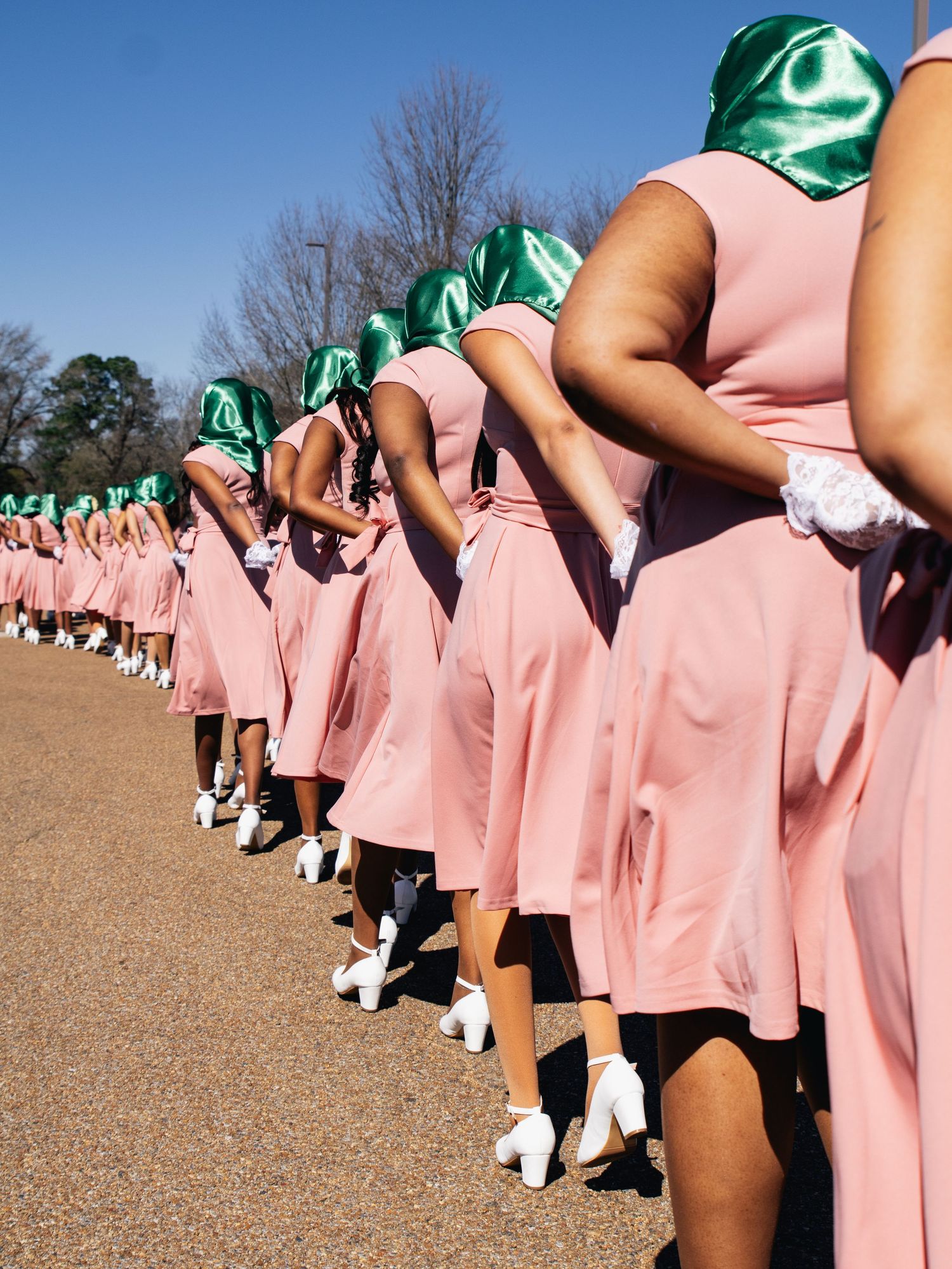Line of people in pink dresses and green accessories standing outdoors.