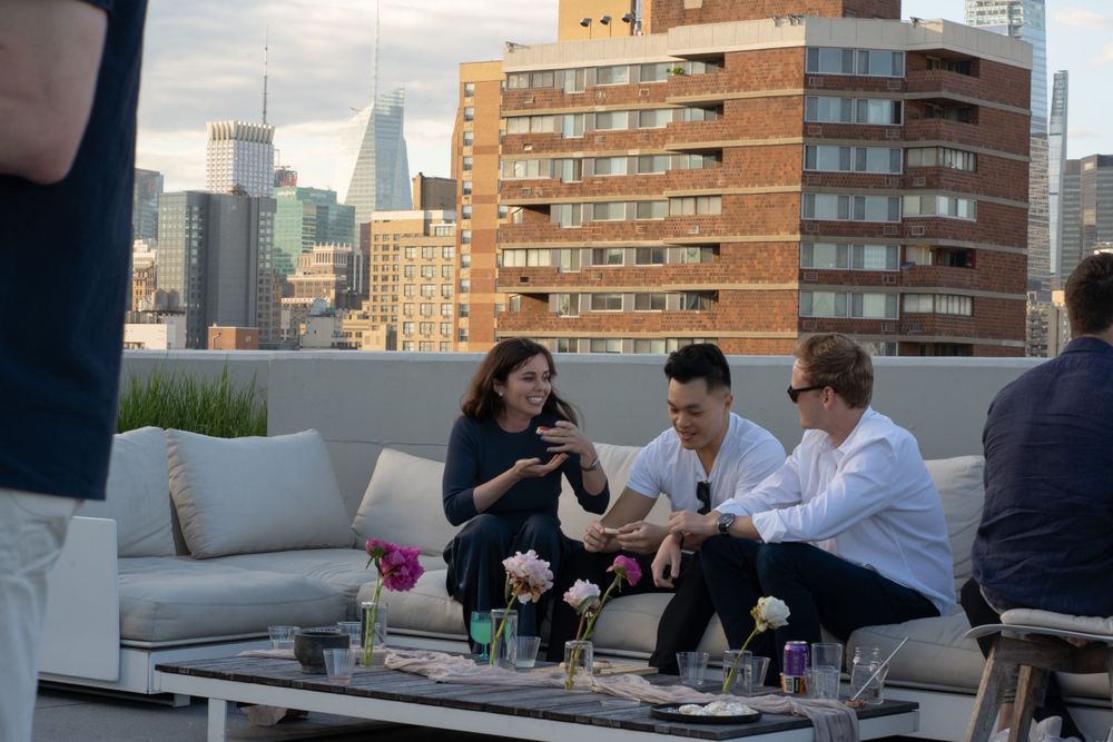 Group chatting on comfortable patio furniture at NYC rooftop party overlooking urban skyline.