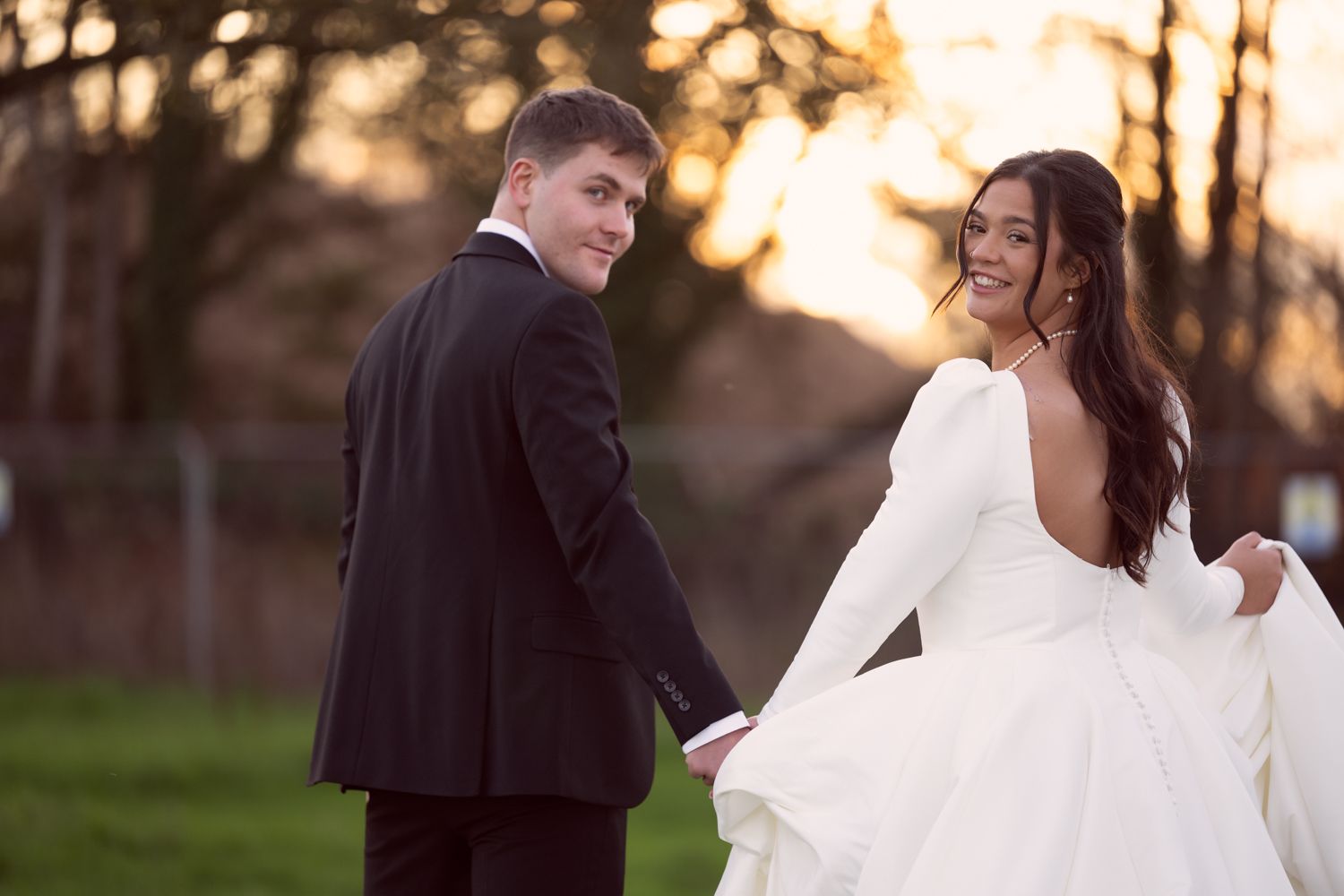 A couple in wedding attire smiles at each other during sunset in a grassy field with trees in the background.
