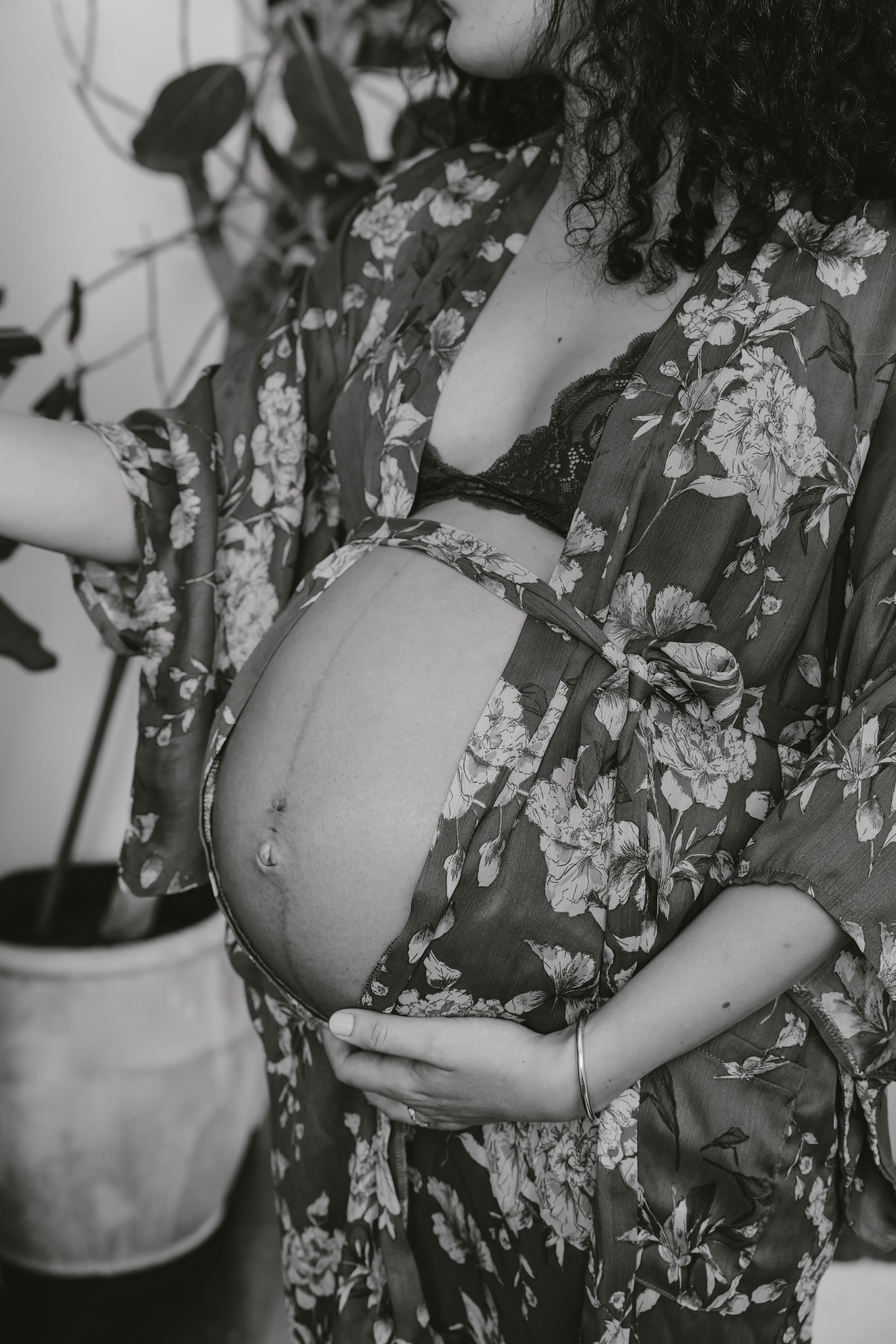 Black and white photo of a pregnant belly in a floral robe with hands cradling the bump near a potted plant.