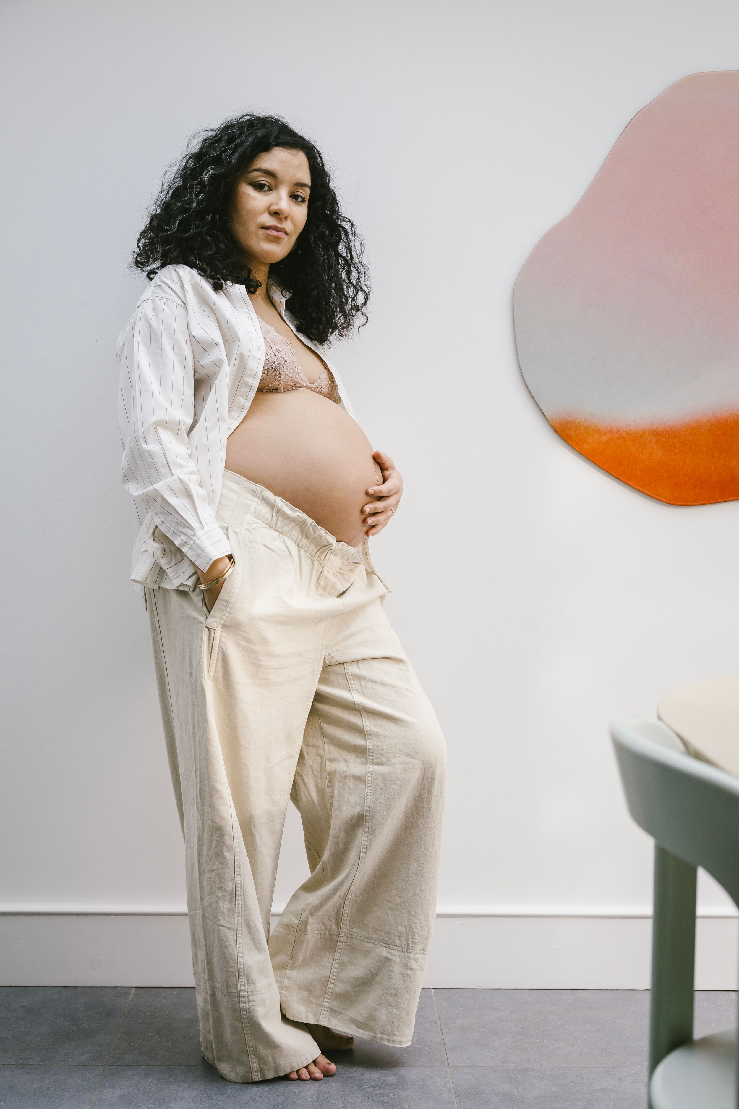A person in cream-colored loungewear and white shirt poses artistically against a minimalist white wall with orange accents.