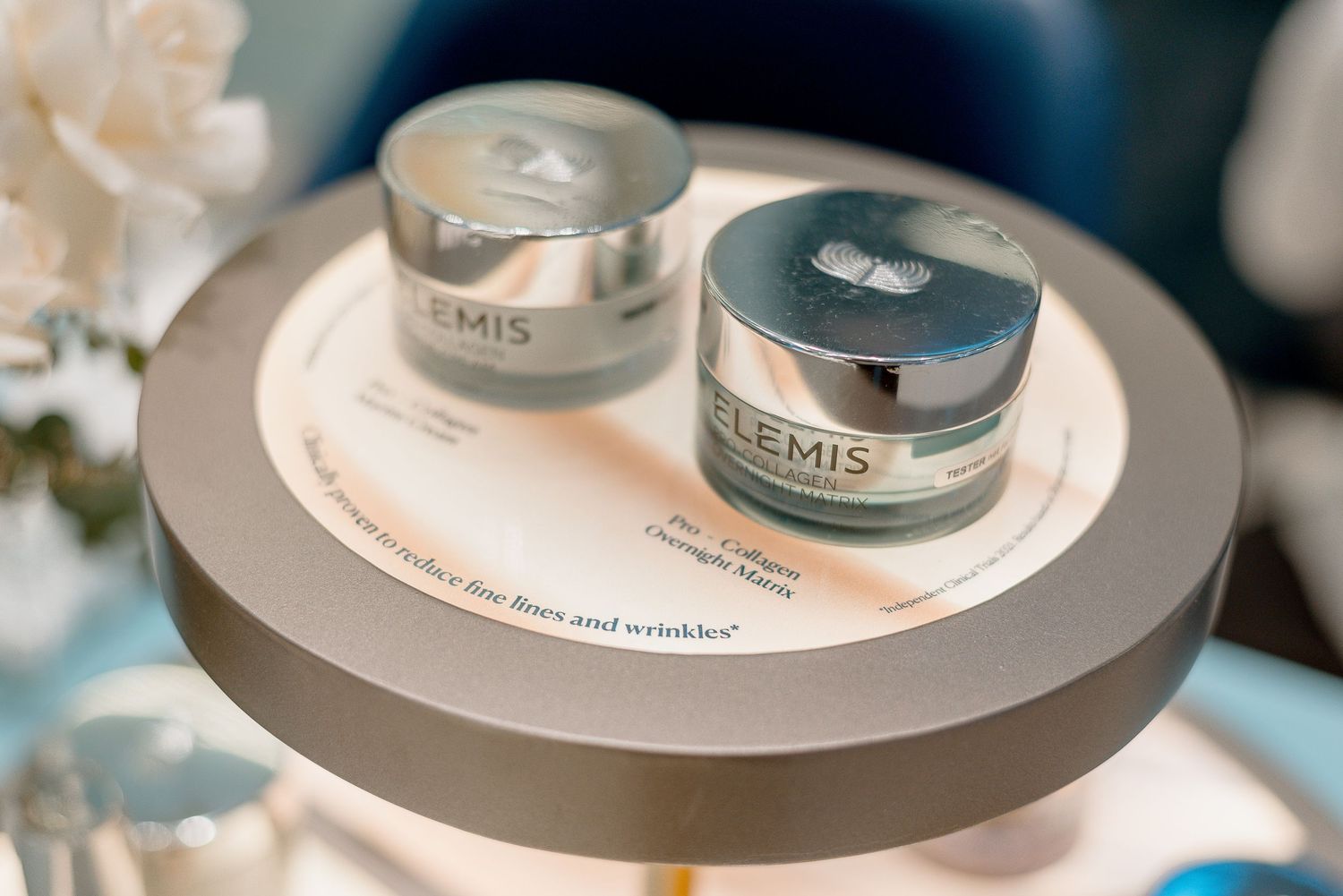 Two Elemis skincare product jars displayed on a circular platform with soft lighting and floral decor.