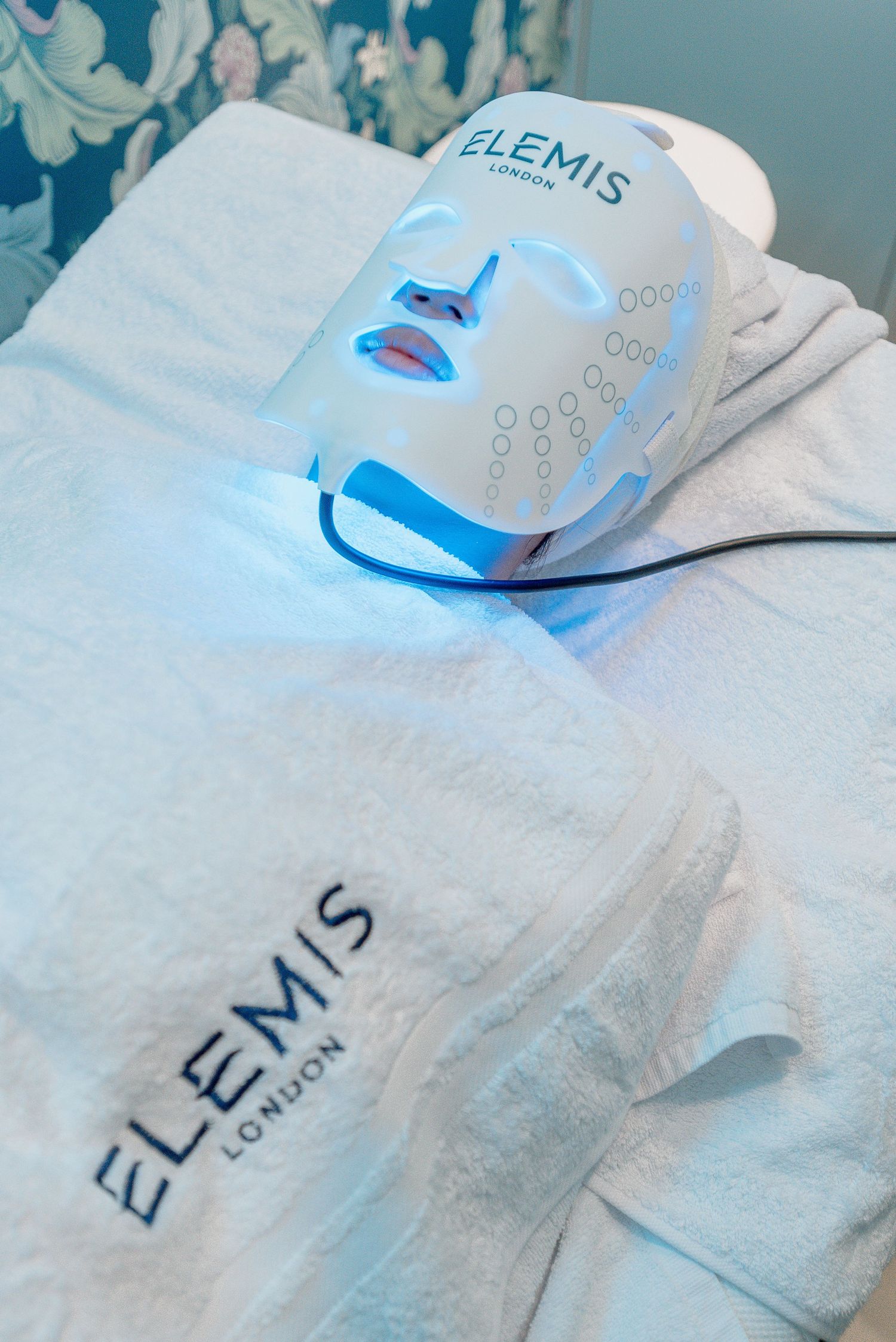 Blue LED light treatment device illuminates white towels and Elemis branded spa products during a skincare treatment session.