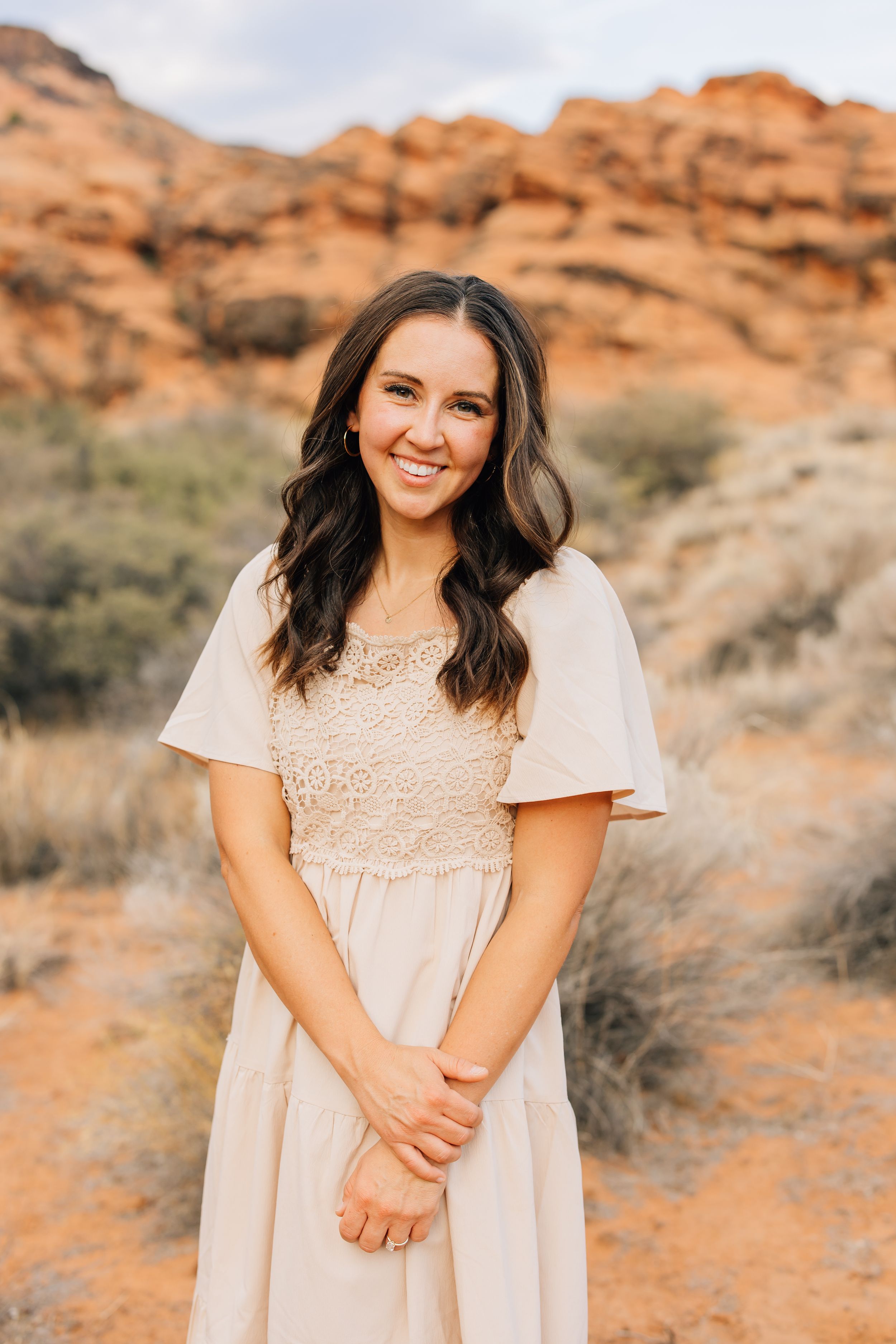 A person in a light beige dress with flutter sleeves poses against red rock formations in a desert landscape.