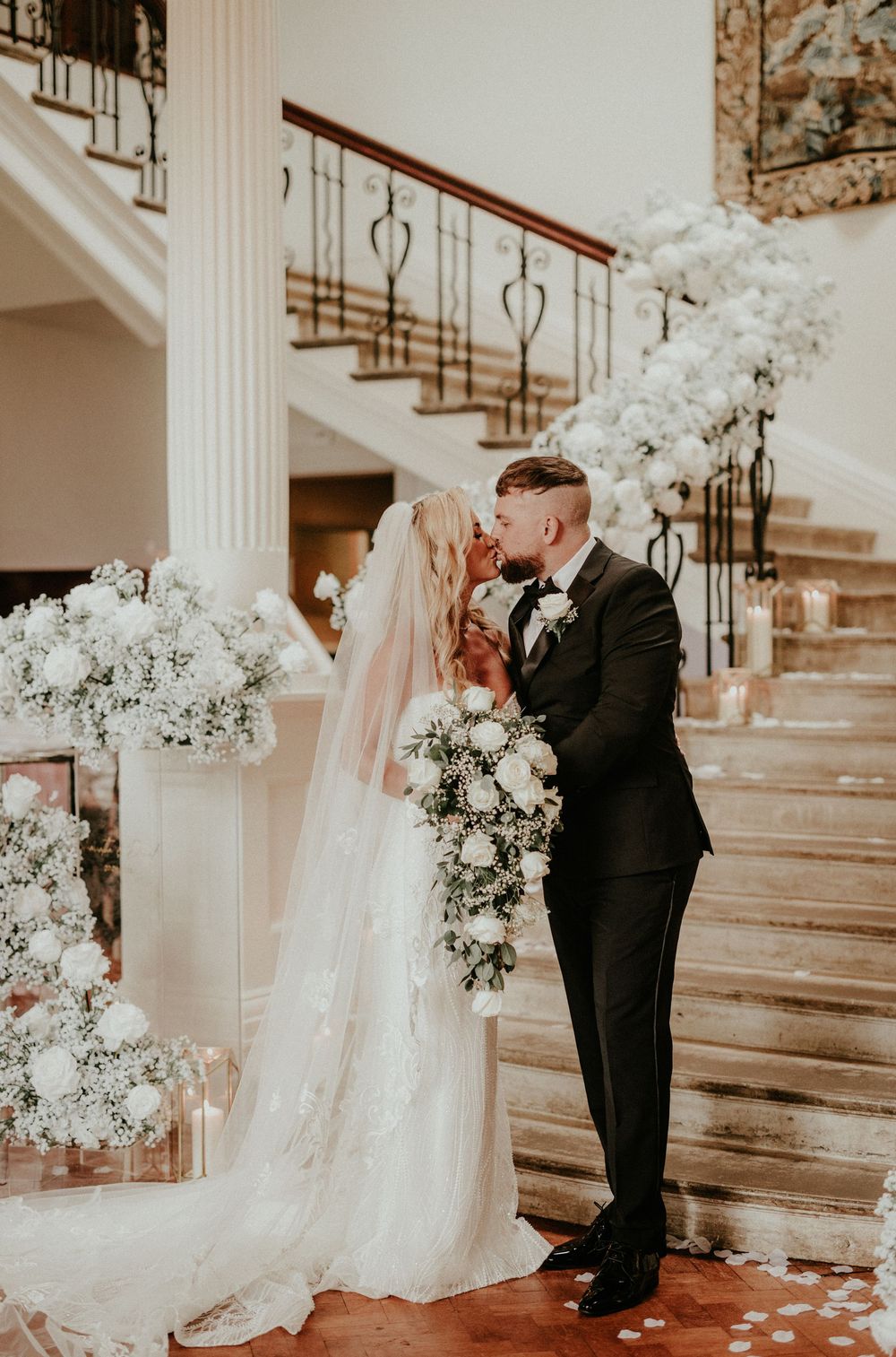 A tender first look moment between newlyweds on an elegant staircase adorned with white floral arrangements.