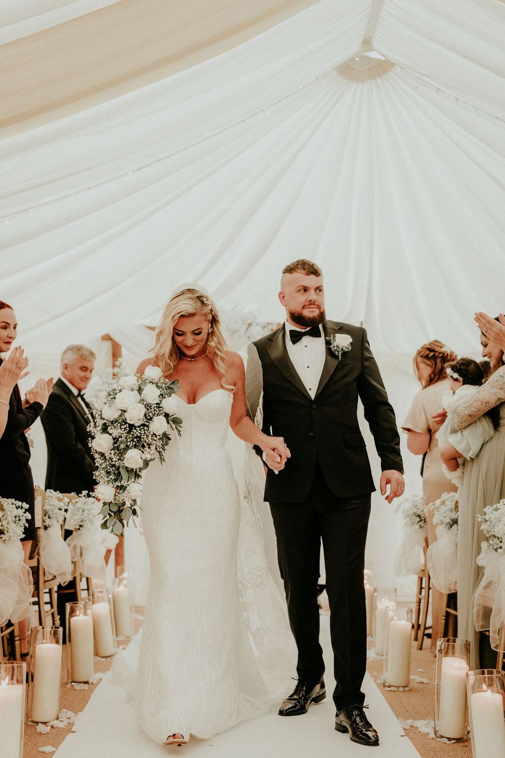 Wedding party in formal attire stands in elegant white tent with bridesmaids in light dresses and groomsmen in black tuxedos.