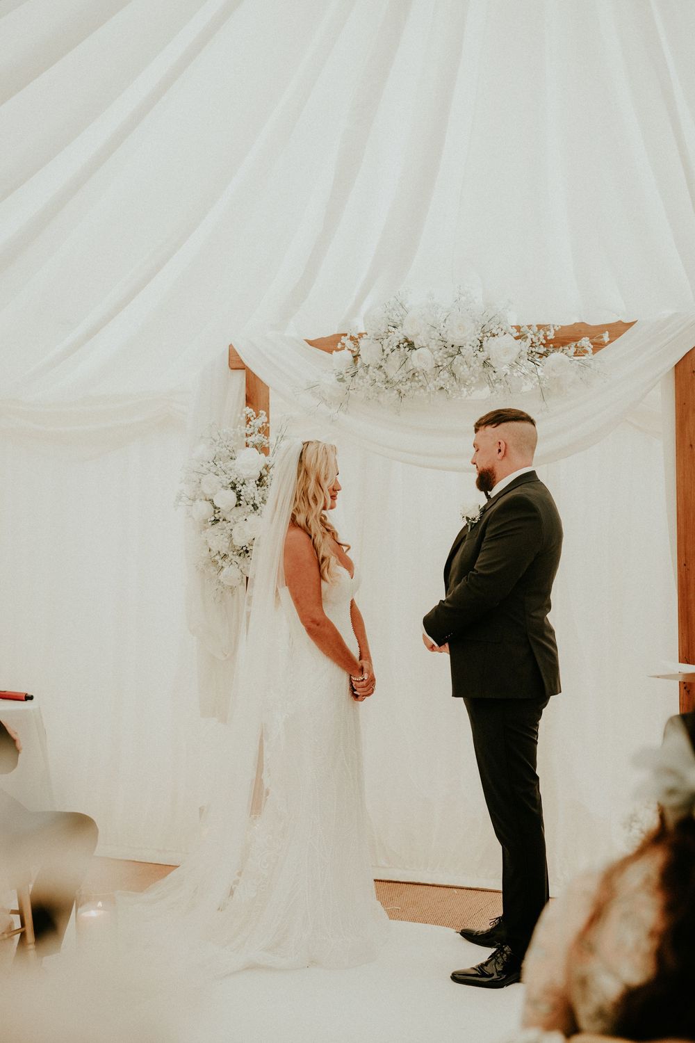 A wedding ceremony in a white draped room with a couple standing before floral arrangements.