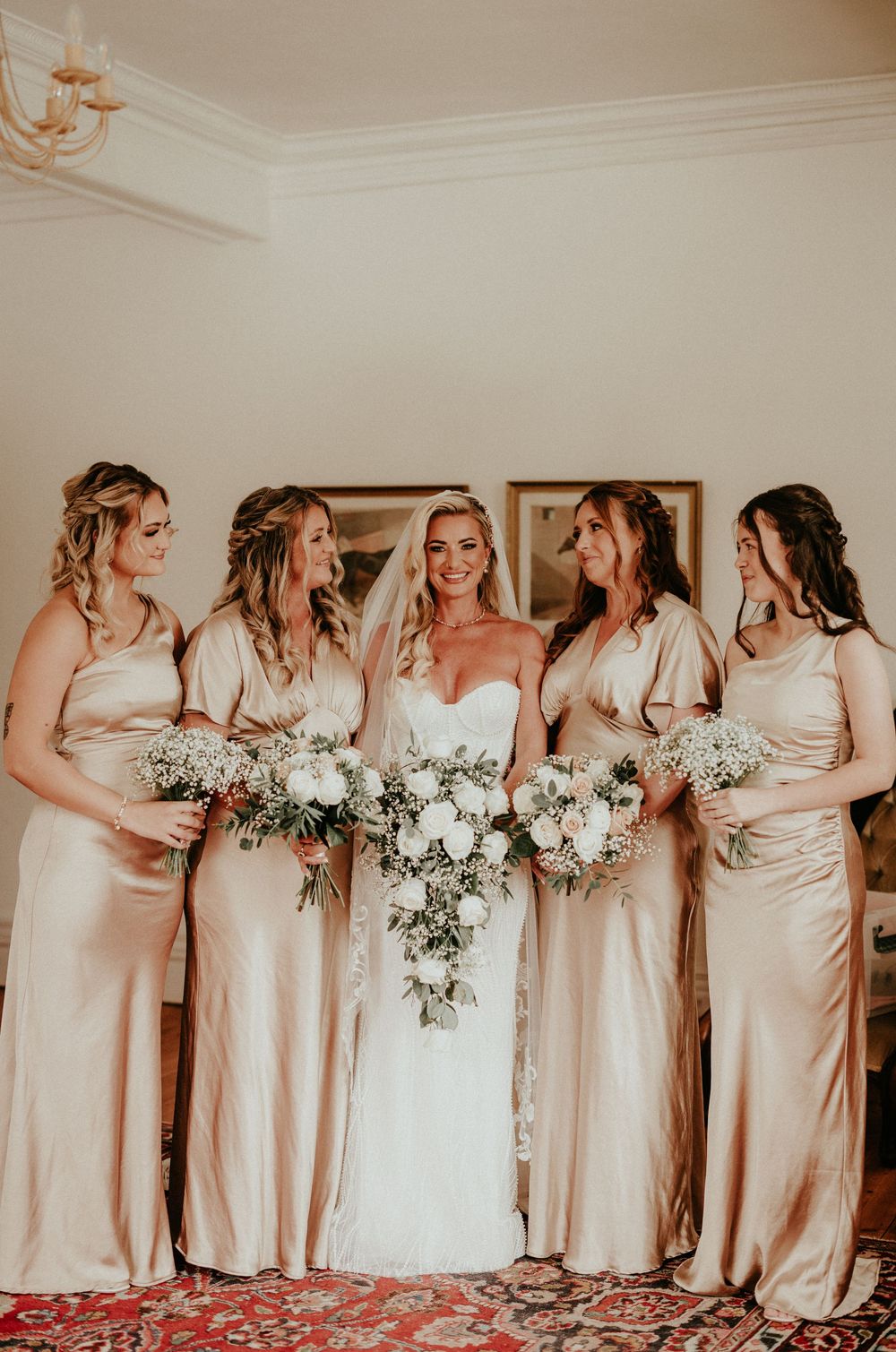 Bridesmaids in champagne satin dresses stand in a row holding white floral bouquets at an elegant wedding.