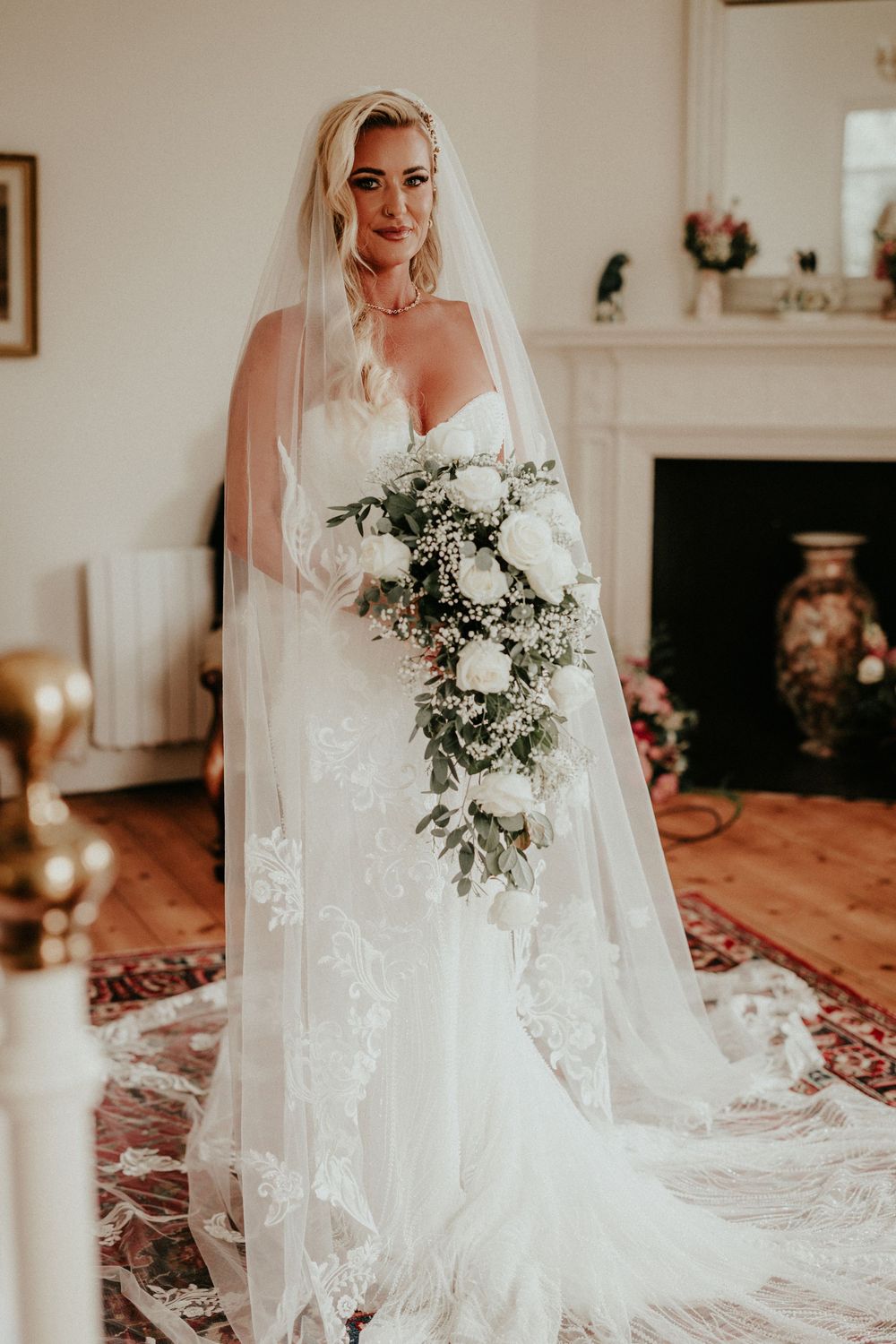 A stunning bride in a white lace gown holds an elegant white and green bouquet against a light interior backdrop.