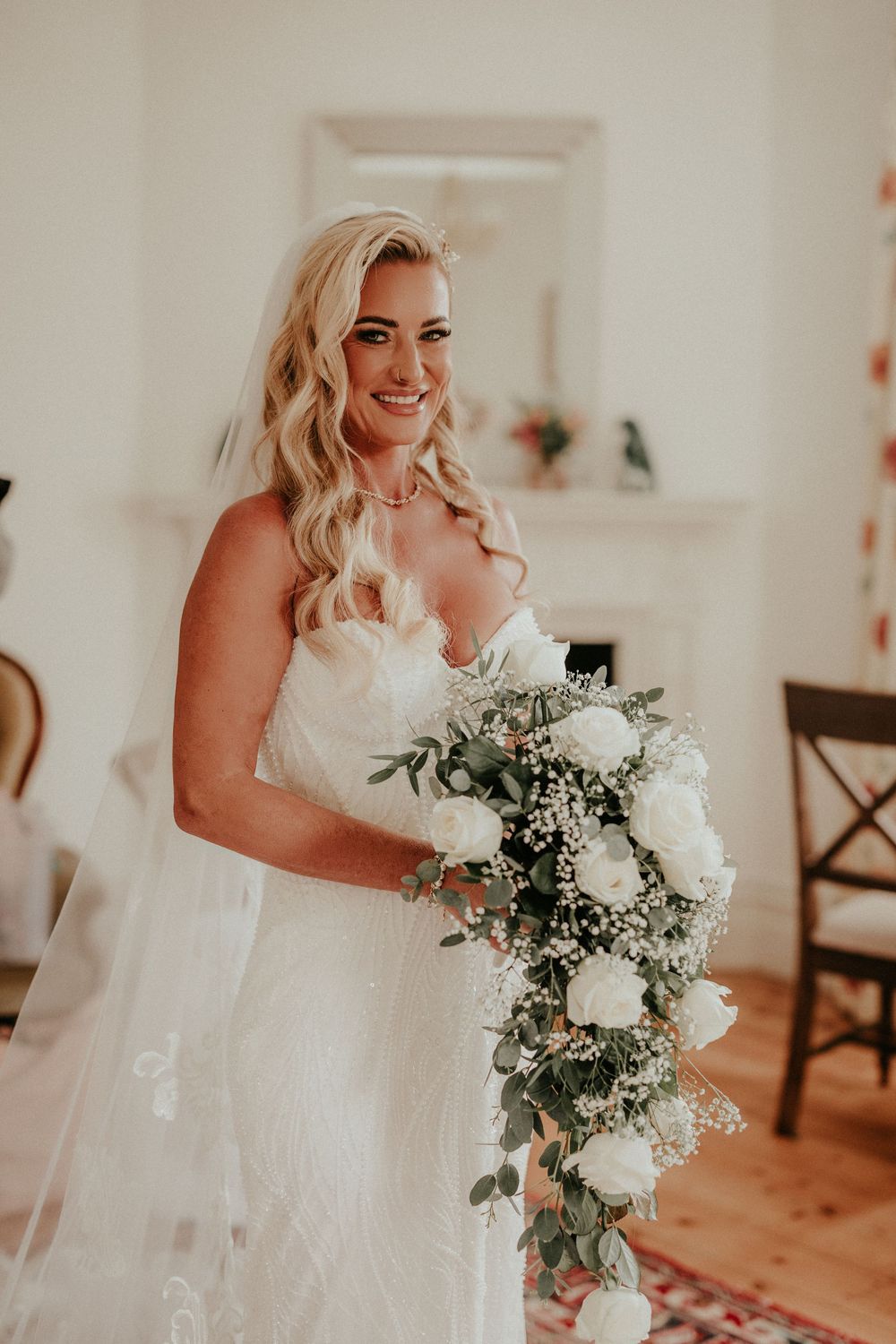 A bride in a white wedding dress holds a beautiful bouquet of white flowers and greenery in a light-filled room.