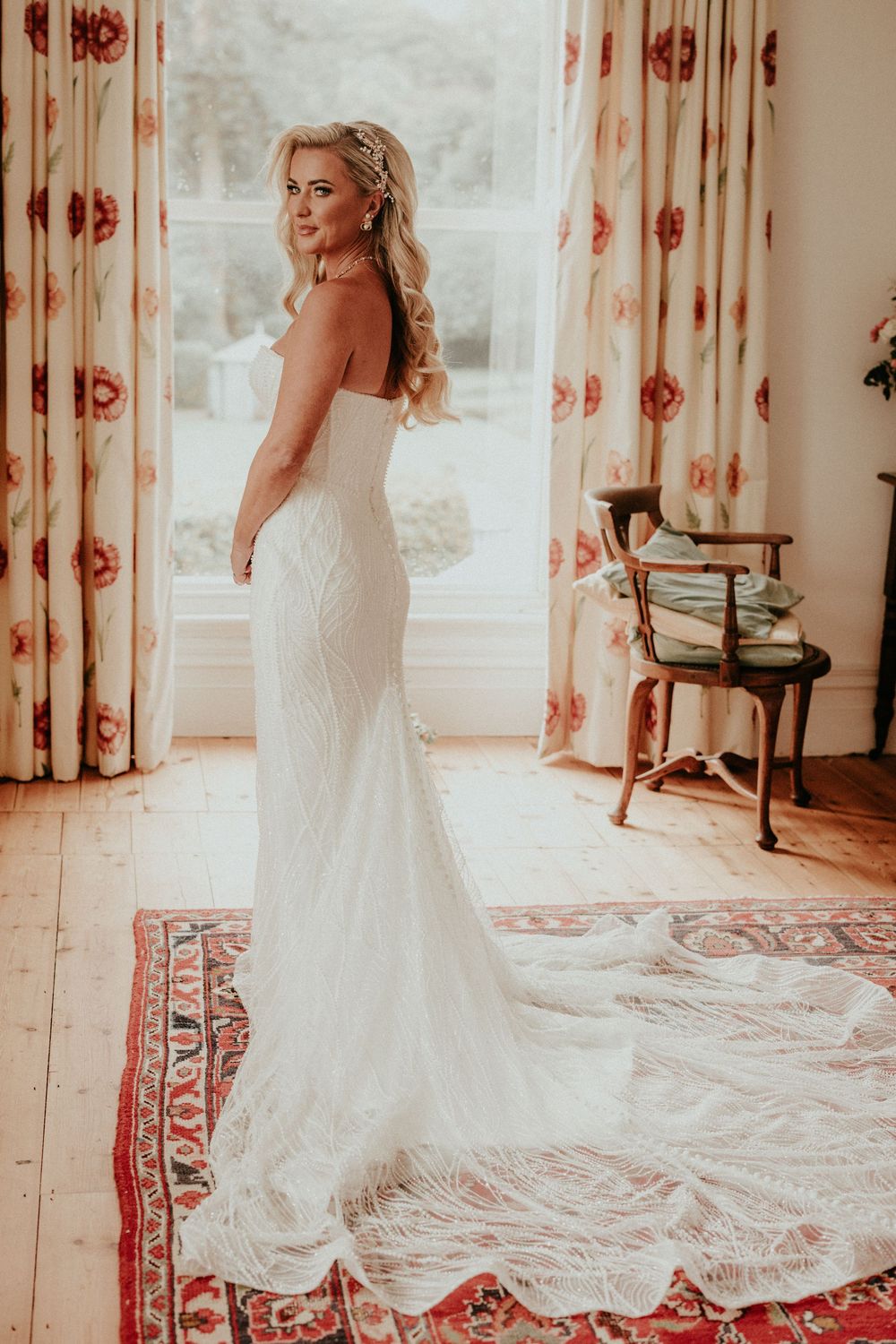 A stunning white lace wedding gown with a long train is showcased in a sunlit room with floral curtains.