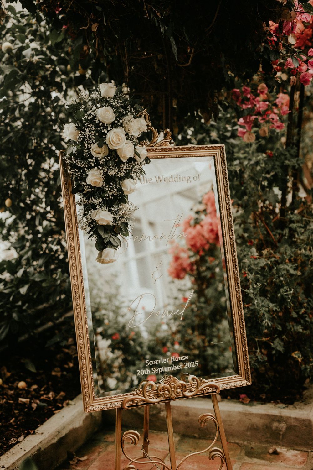 A vintage gold-framed mirror with ornate details sits among blooming flowers and greenery in an outdoor garden setting.