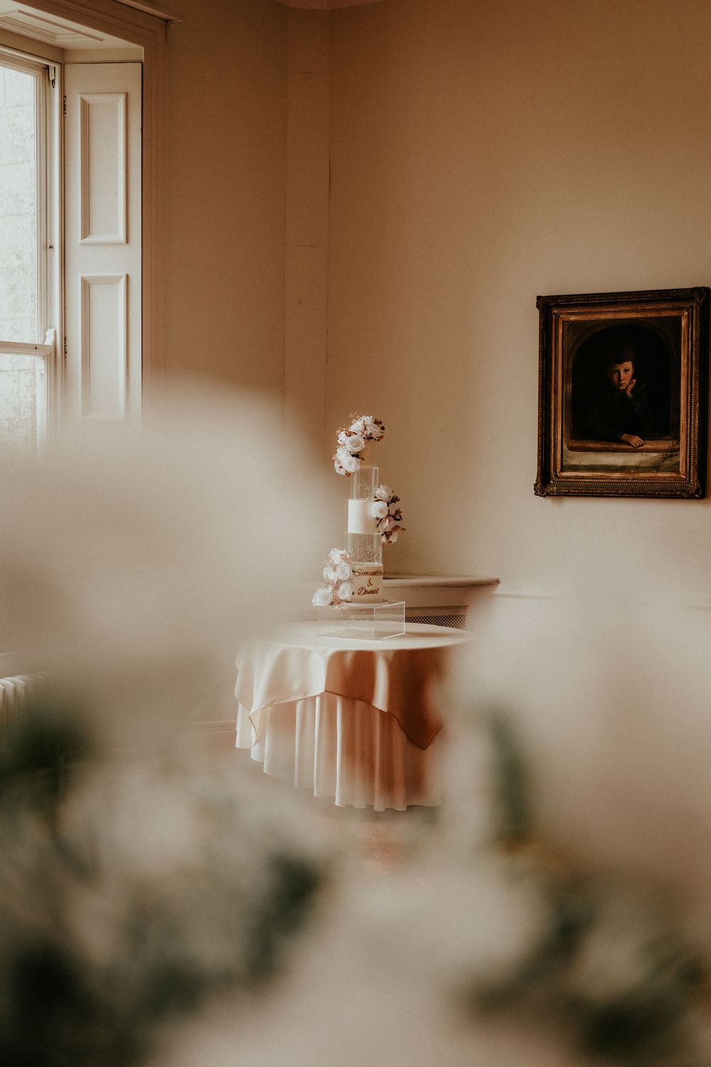 Soft sunlight streams through window onto round table with white cloth and flowers, with vintage portrait on wall.