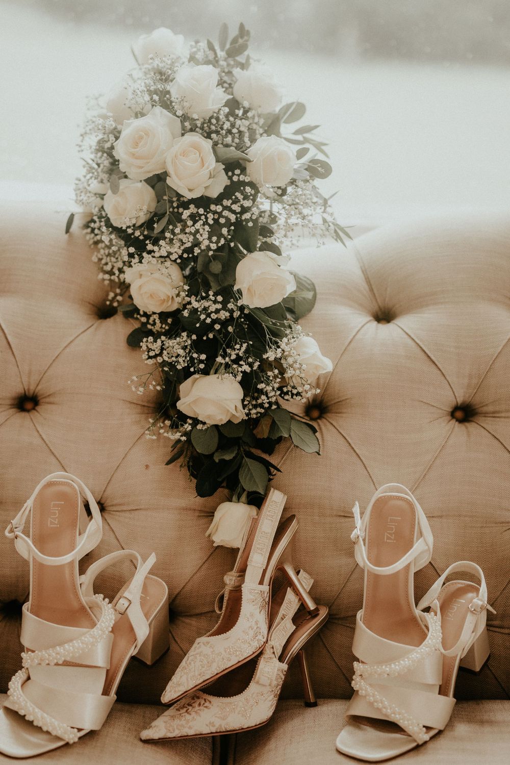 White bridal bouquet with roses and baby's breath rests on beige tufted couch beside three elegant wedding shoes.