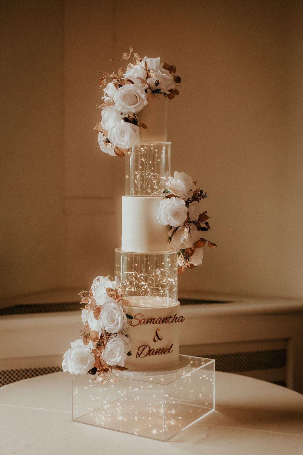 An elegant white wedding cake decorated with roses and fairy lights sits on a crystal stand. Cornwall wedding photography scorrier house